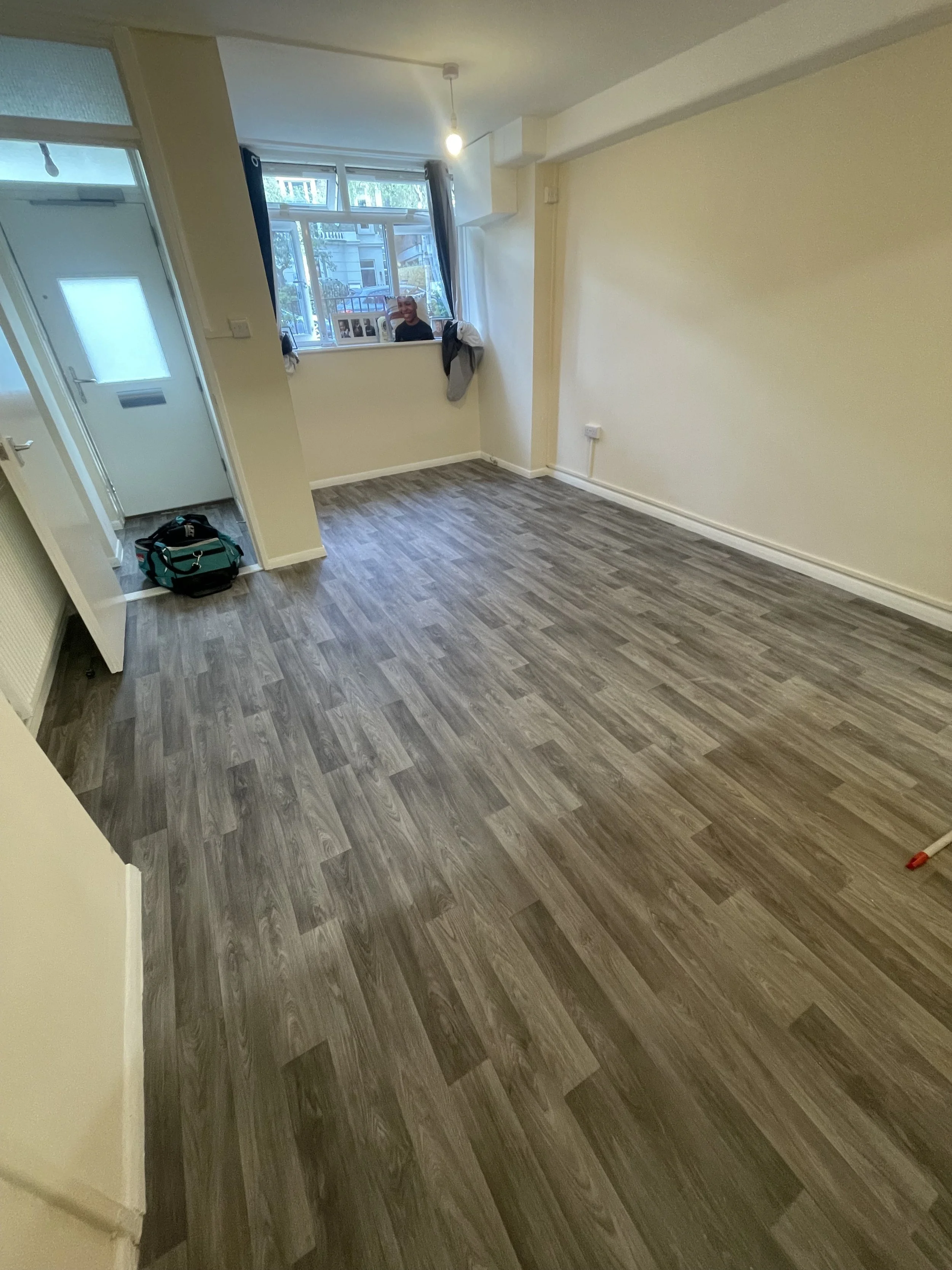 Empty living room with wood floor, beige walls, a window with blue curtains, and a person sitting on the windowsill. There is a backpack on the floor near the door, and a laundry marker in the corner.