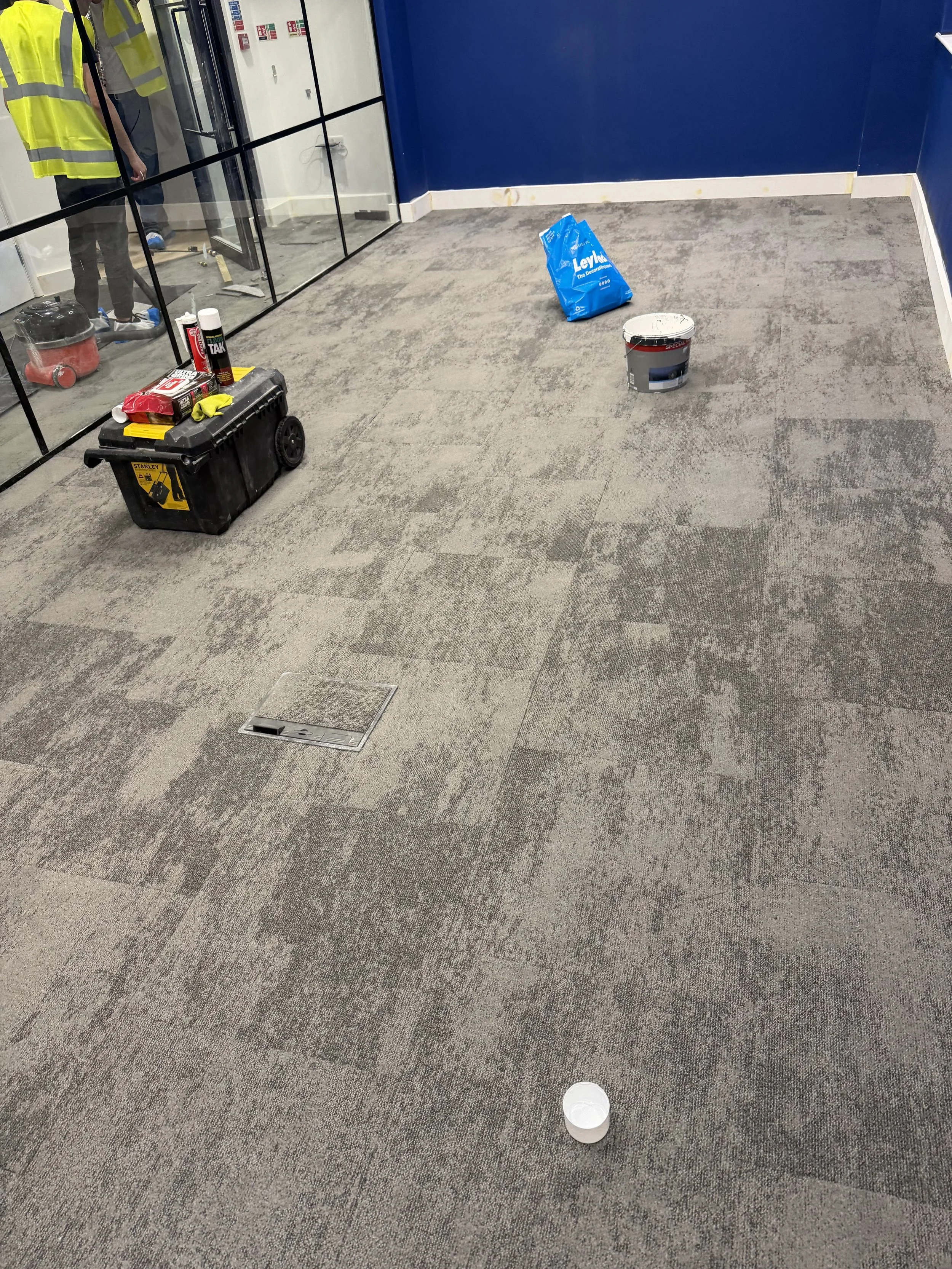 Empty room with carpet flooring, construction tools, a blue bag, paint bucket, and a small white cup on the floor.