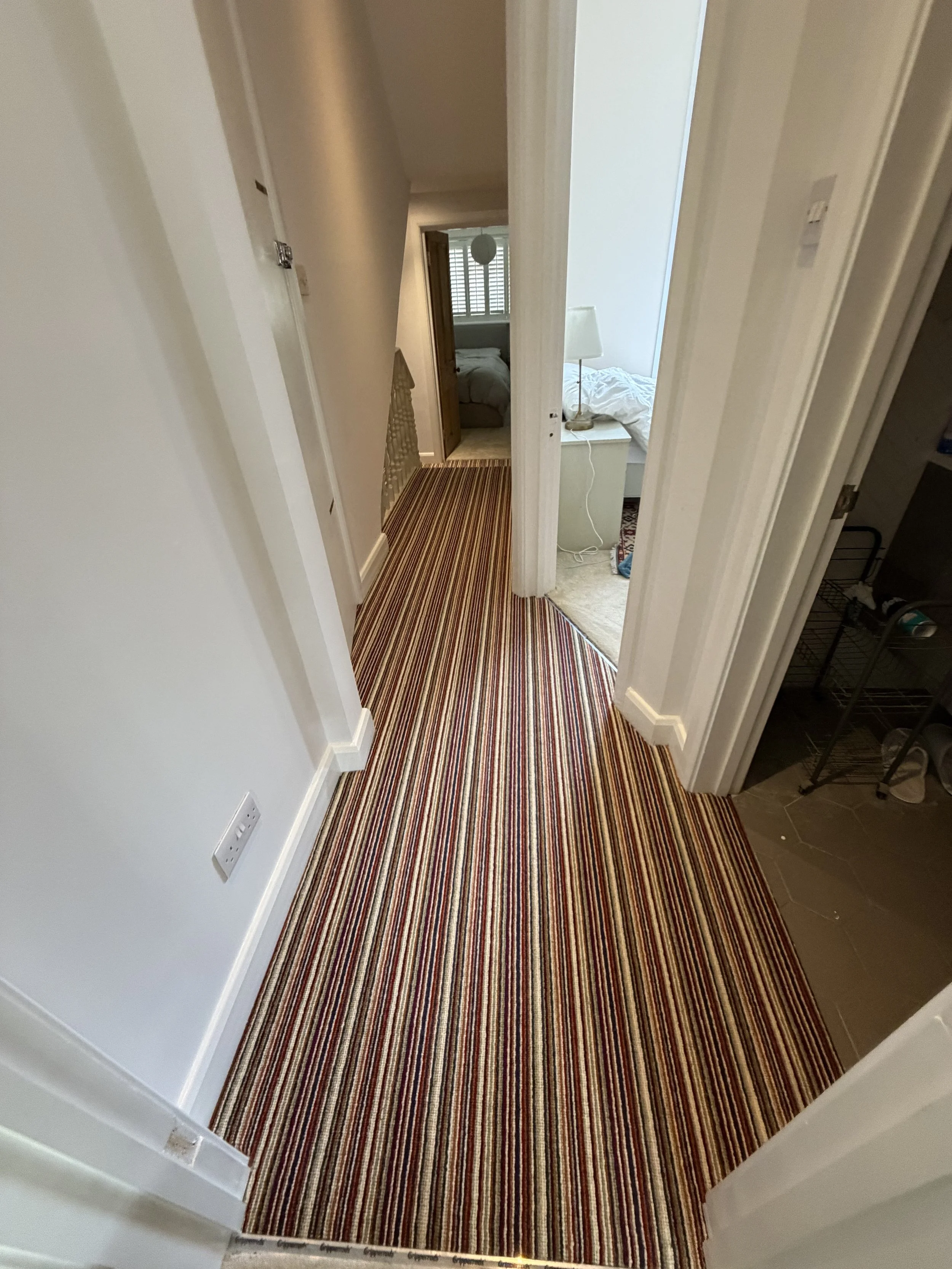 View of a hallway in a home with a striped carpet, leading to bedrooms with visible beds and furniture.