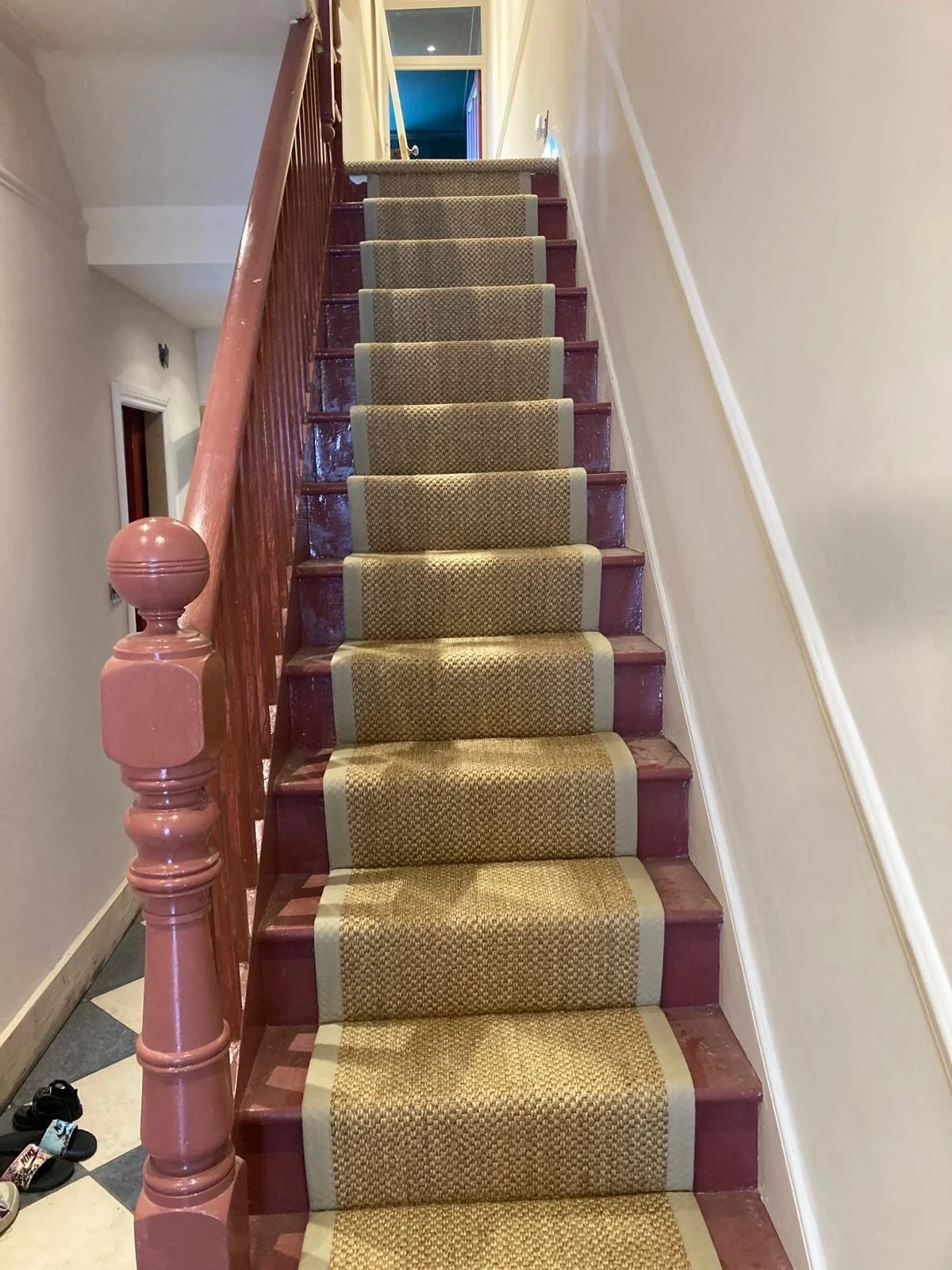 Indoor staircase with a beige carpet runner, reddish-brown wooden handrail and steps, walls painted in light cream, and shoes near the bottom of the stairs.