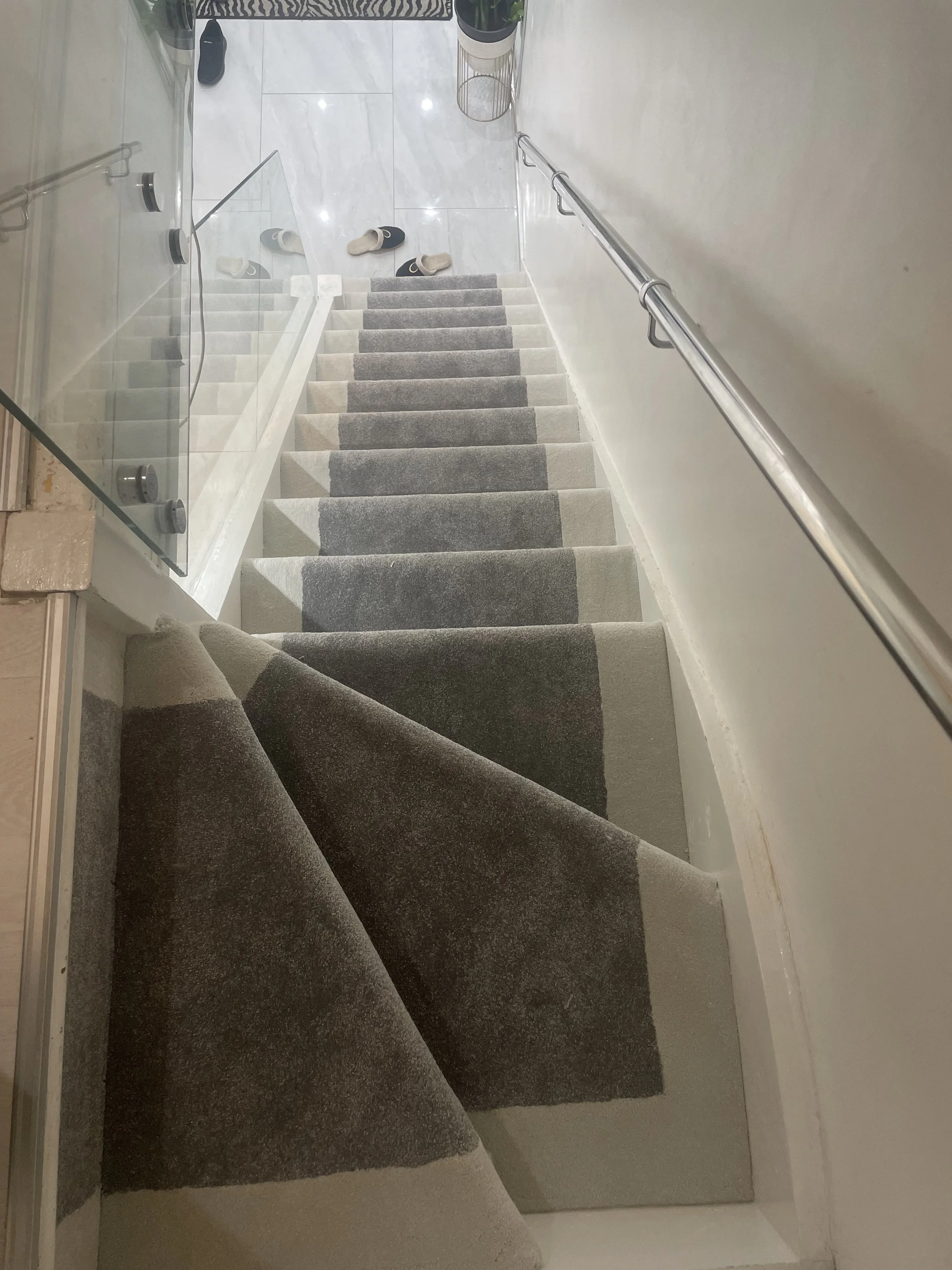 Indoor staircase with beige and gray striped carpet runner, handrail on the right, slippers at the top, glass barrier on the left, and a tiled floor at the top of the stairs.