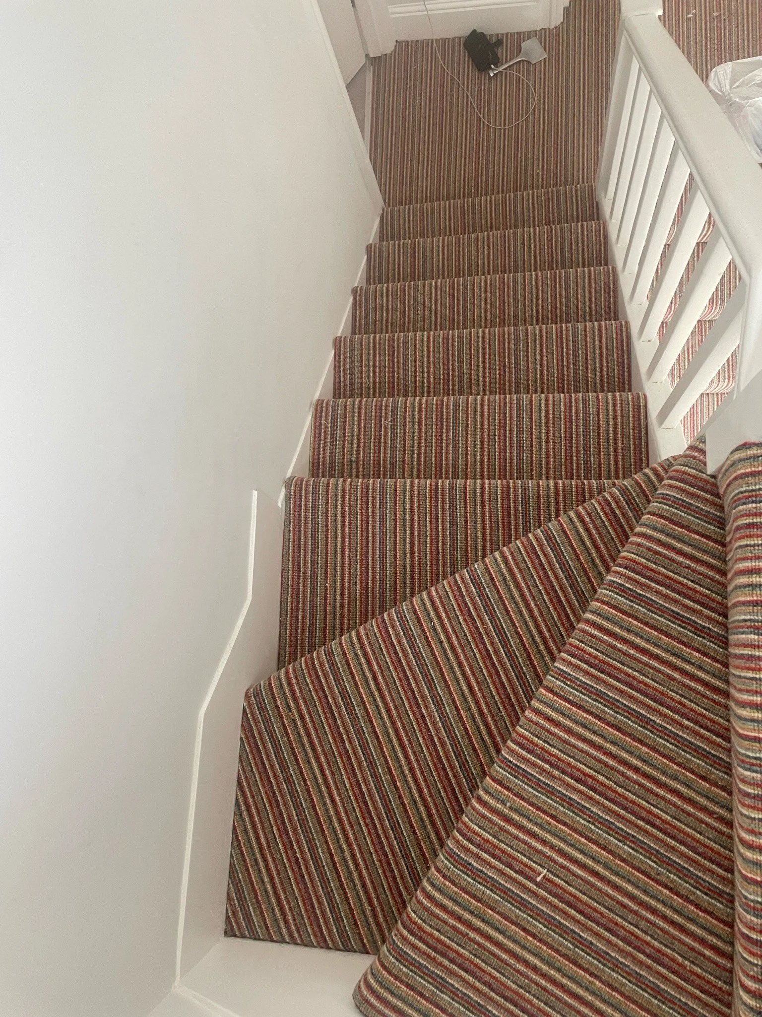 Overhead view of a staircase with striped carpet treads, white railings, and a wall on the left side.