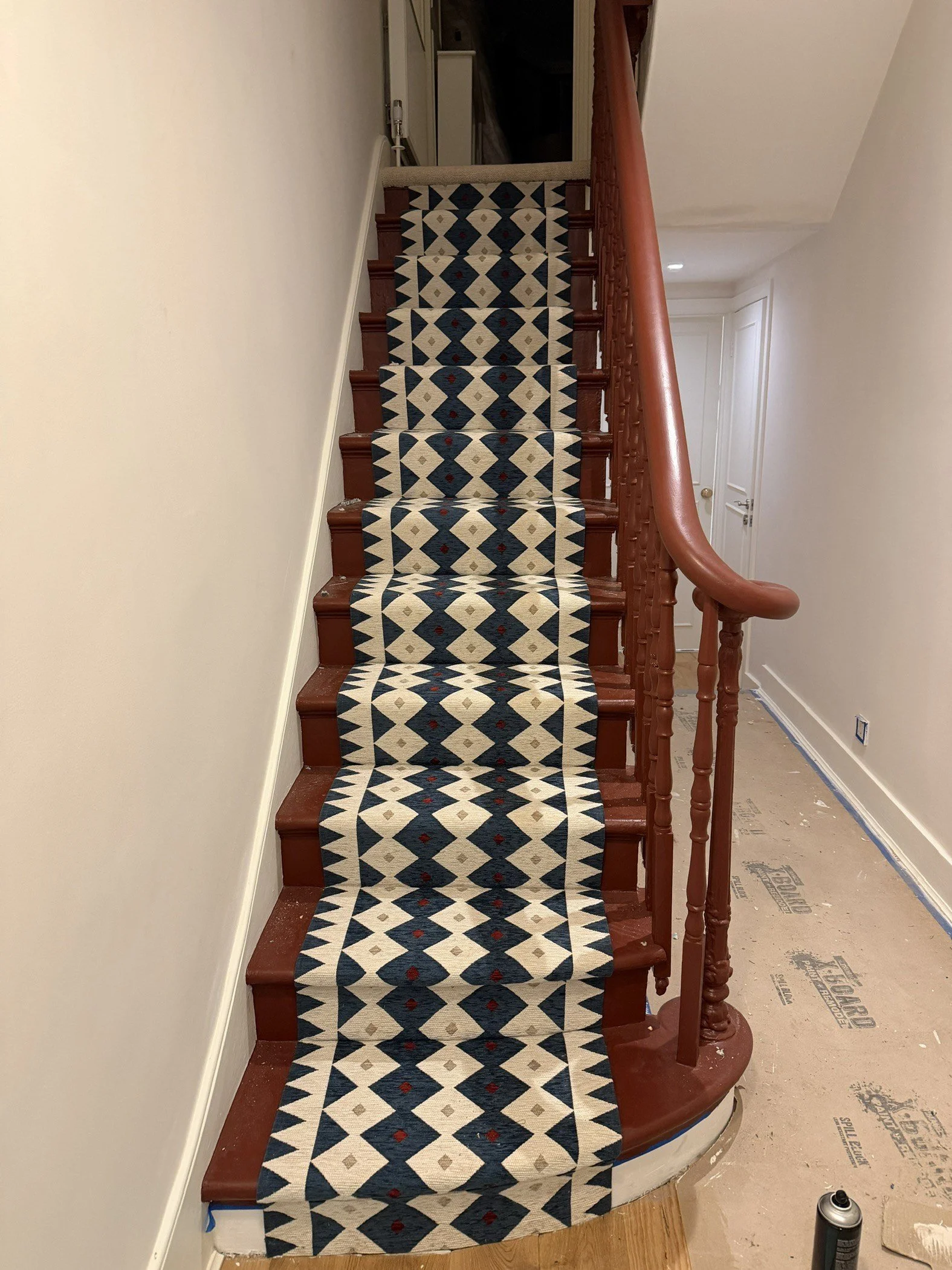 A staircase with a patterned runner carpet. The carpet features a geometric design in navy blue, beige, and small red accents. The staircase has a reddish-brown wooden handrail and spindles.