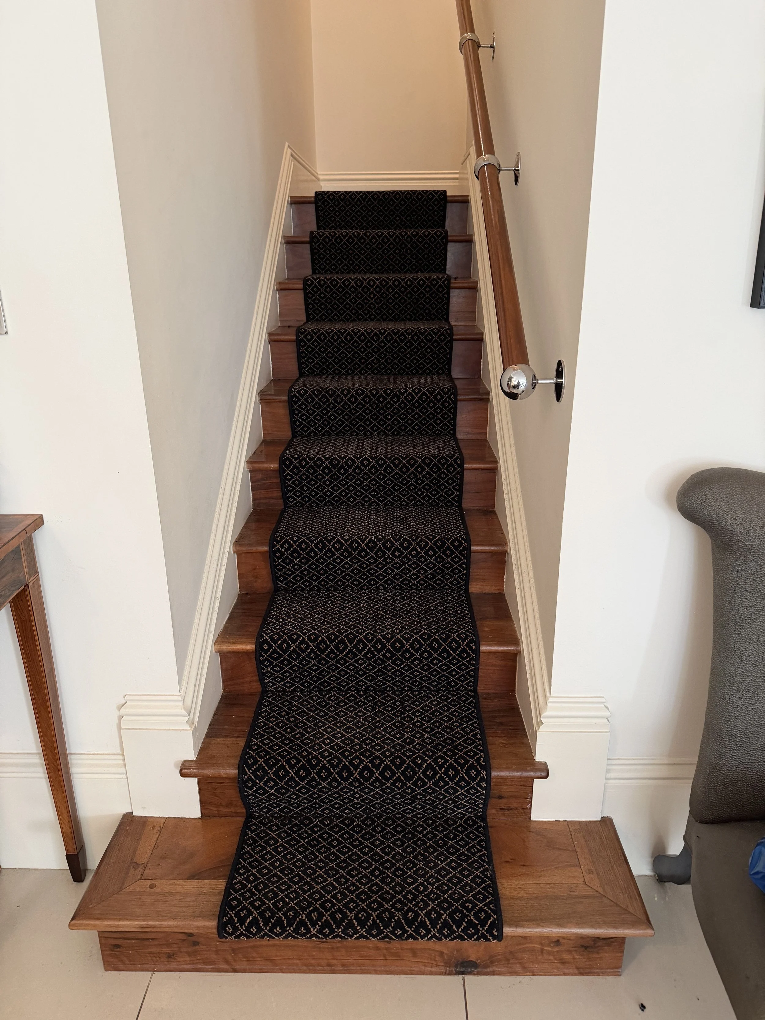 Wooden staircase with a black patterned carpet runner, white walls, and a wooden handrail on the right side.