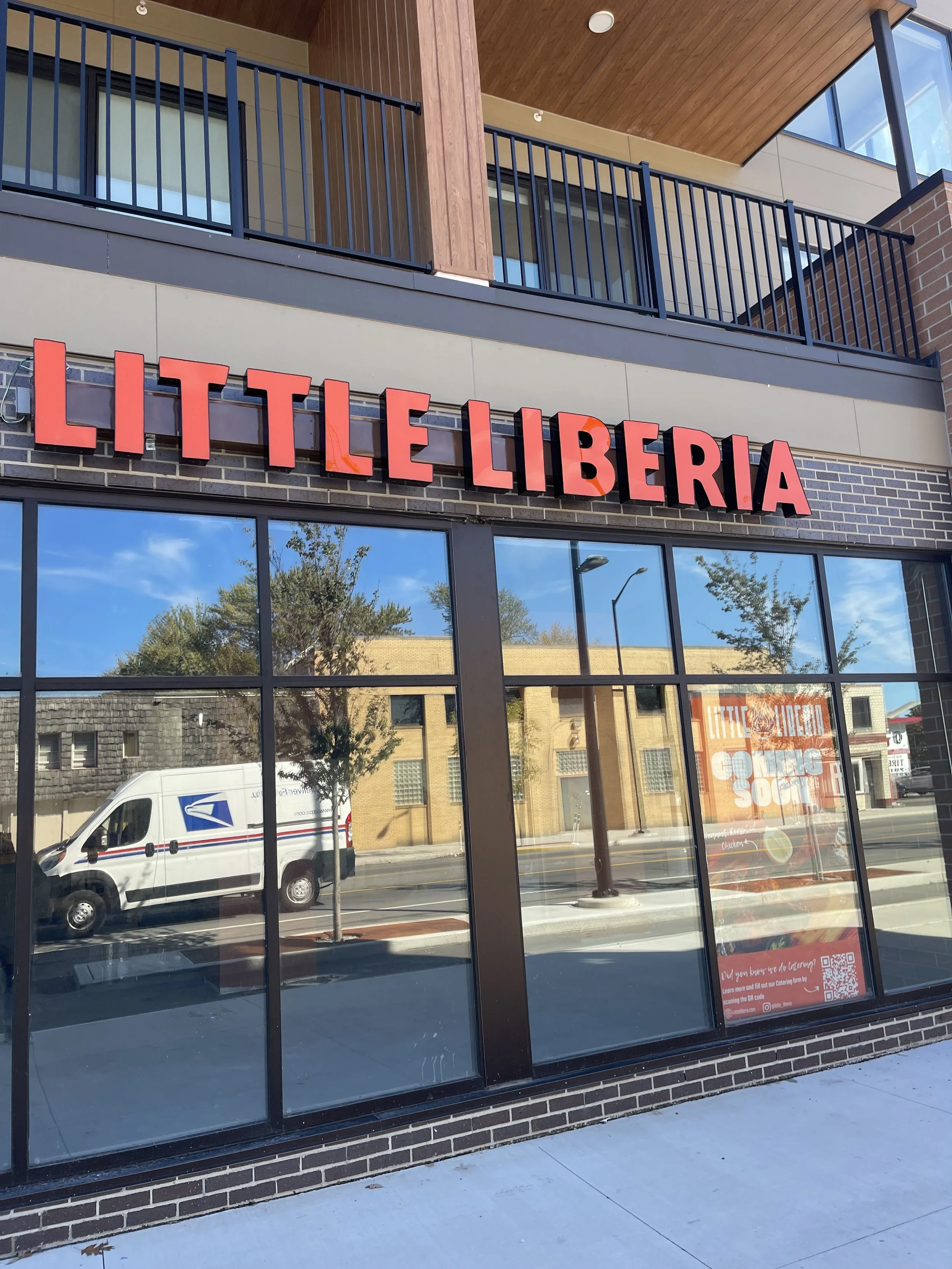 Exterior of a storefront with a sign that reads 'LITTLE LIBERIA' in large red letters. The store has large glass windows reflecting the street scene outside, including trees, buildings, and a USPS postal vehicle.