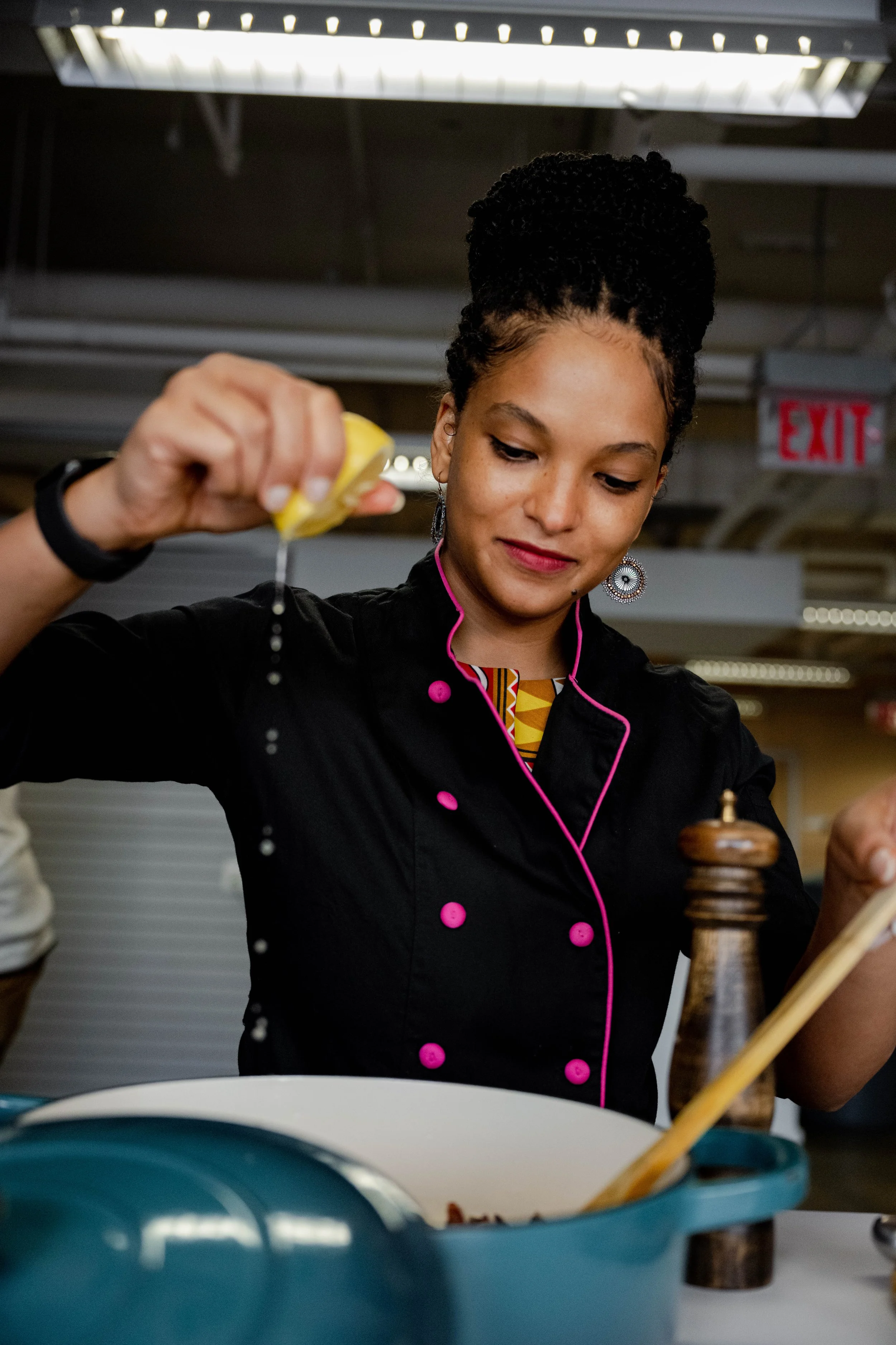 A woman in a black chef's coat with pink piping is squeezing lemon juice into a blue cooking pot. The setting appears to be a professional kitchen, with a pepper grinder and wooden spoon nearby.