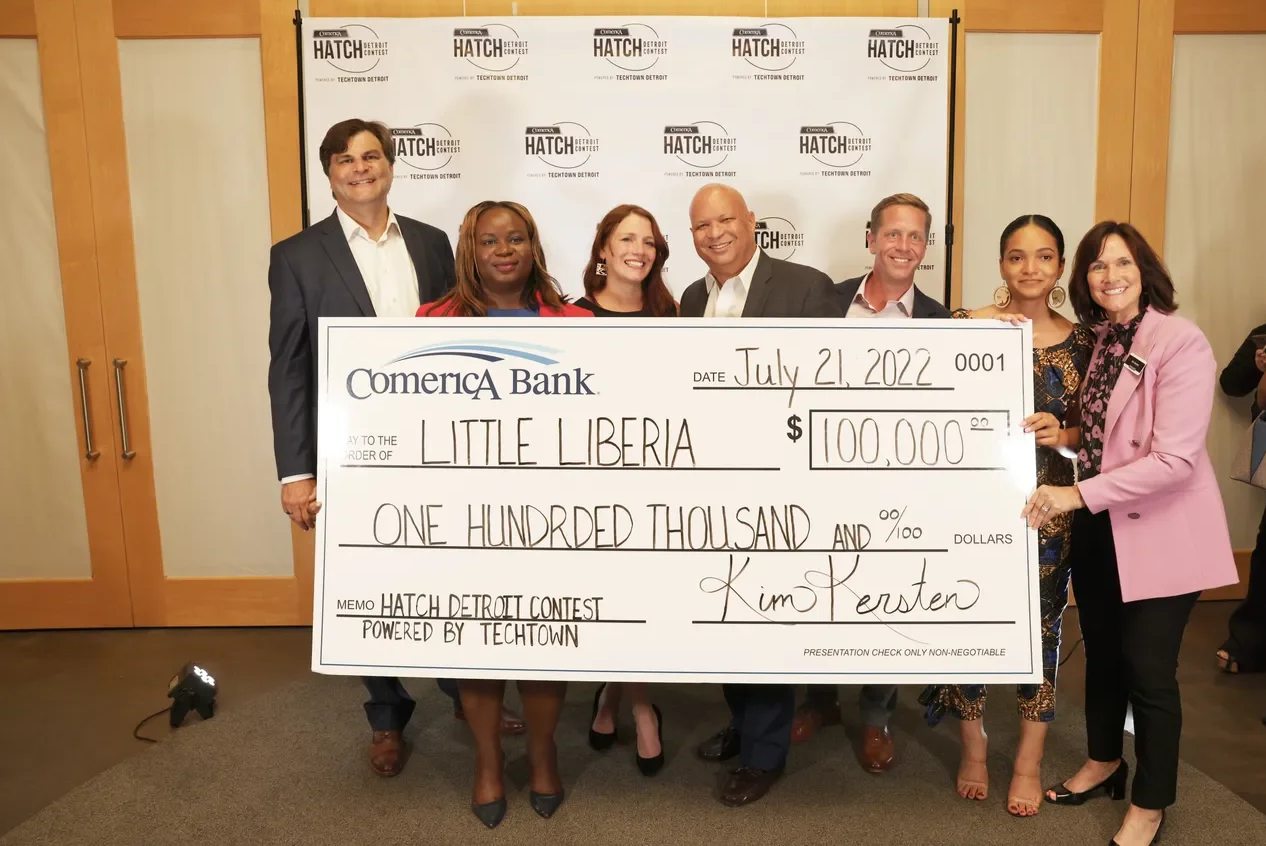 Group of eight people standing in front of a branded backdrop, holding an oversized check for $100,000 made out to Little Liberia, dated July 21, 2022, during a formal event or celebration.