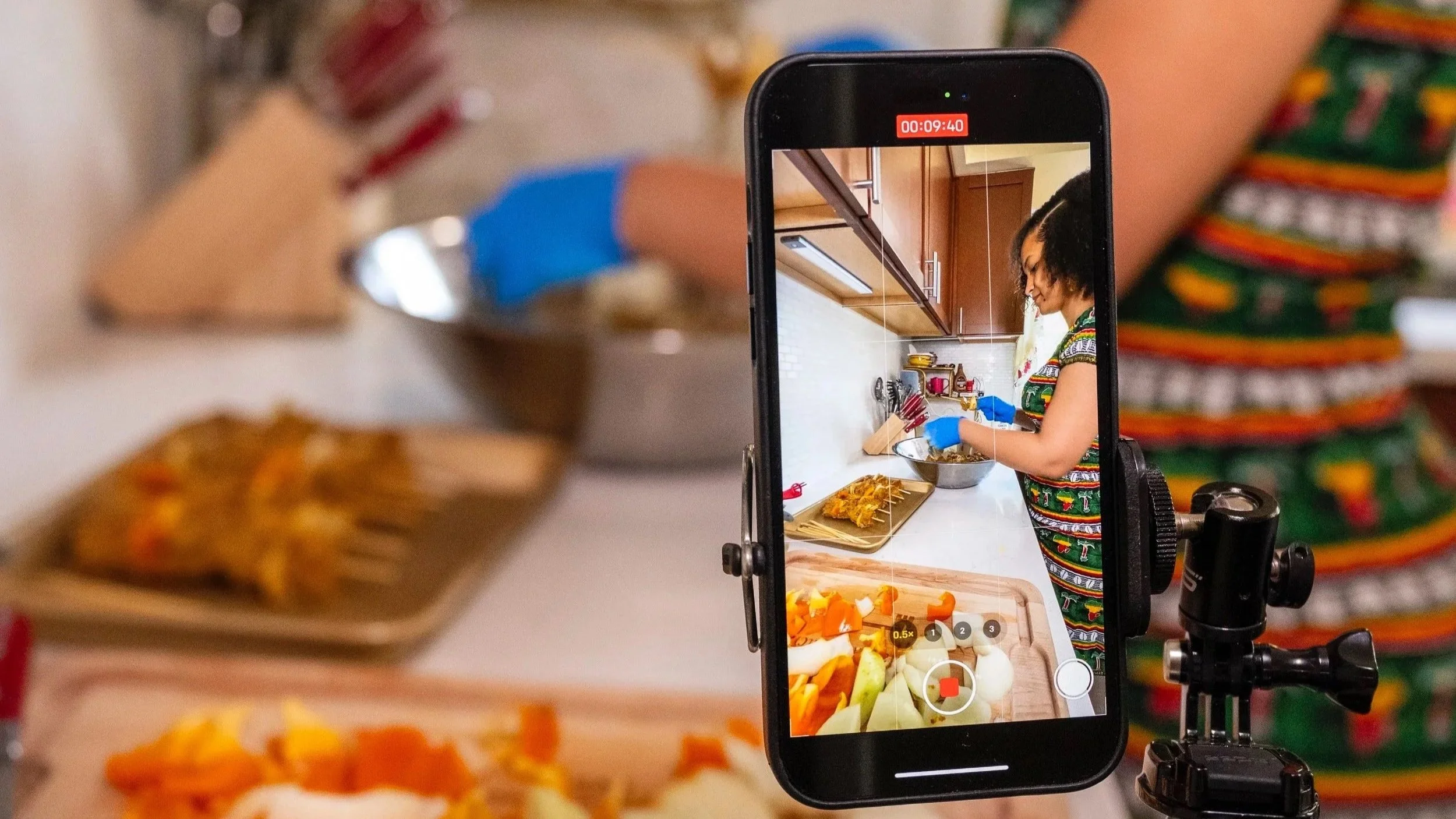 A woman in a colorful dress is filming a cooking process in a kitchen using a smartphone mounted on a tripod. The woman is wearing blue gloves and preparing food with vegetables and other ingredients on a white countertop.
