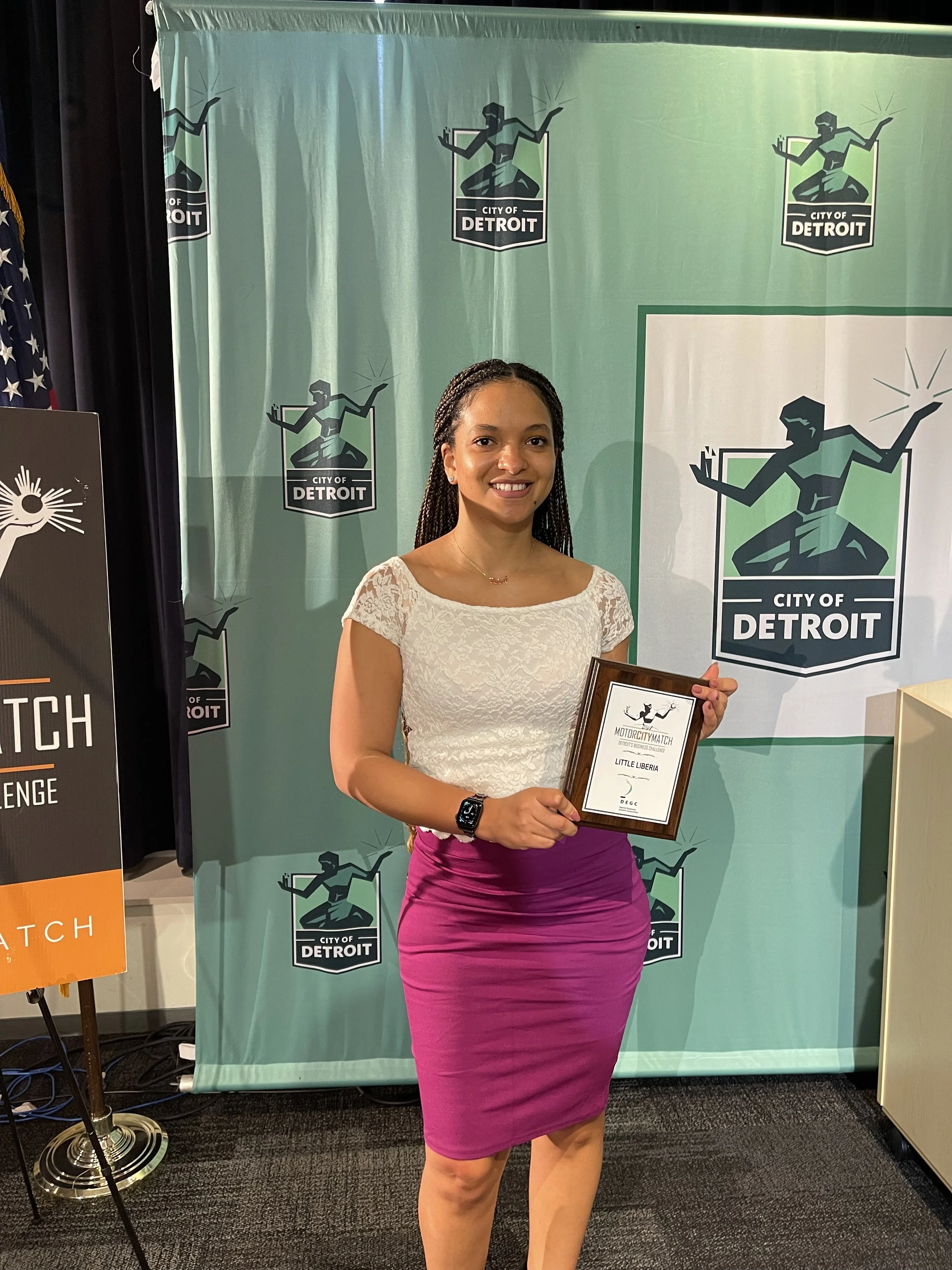 A woman standing and smiling while holding a framed certficate in front of a Green backdrop with the City of Detroit logo. She is dressed in a white lace top and a purple skirt.