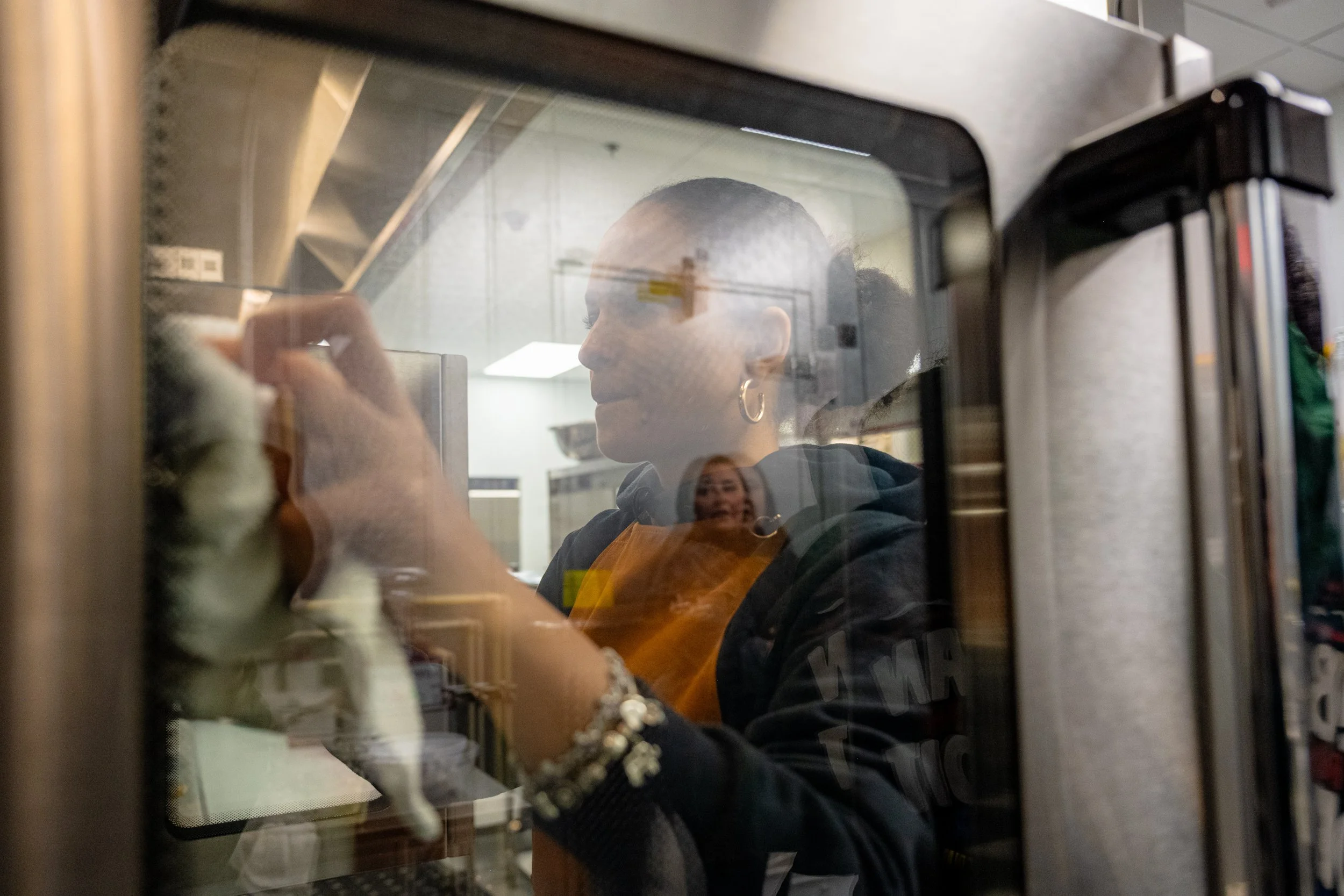 A person with earrings and a bracelet using a microwave oven, viewed through the glass door, with the person holding a spoon and looking inside.