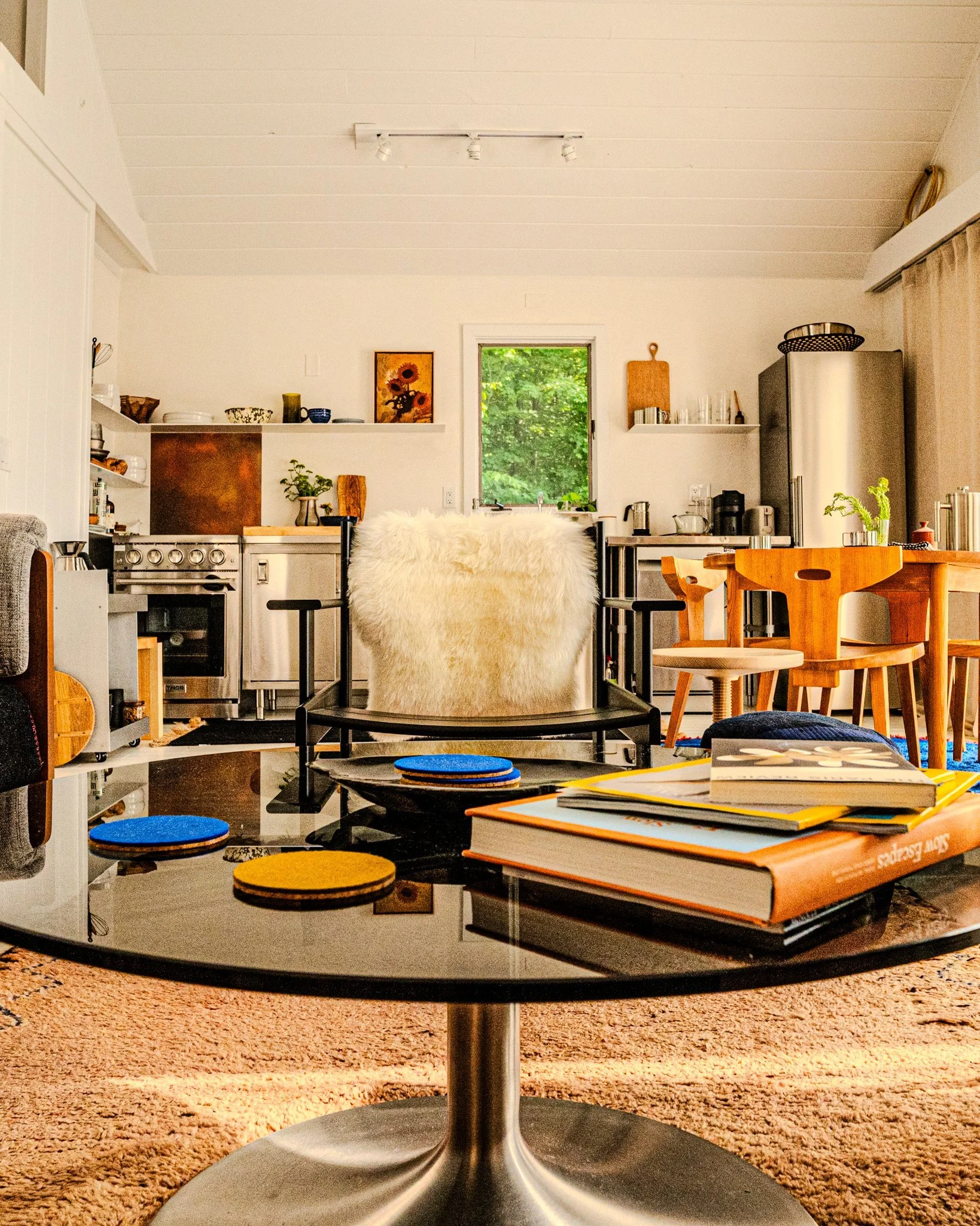 Living room with black coffee table holding books and coasters, kitchen in the background with white walls, wooden dining table, chairs, and window showing greenery outside.