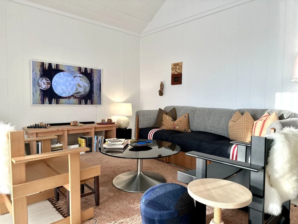 A cozy living room with a gray sectional sofa decorated with colorful pillows, a black round coffee table, a wooden armchair with a furry white throw, and a long wooden sideboard with books and decor. There's a wall-mounted art piece, a lamp, and som