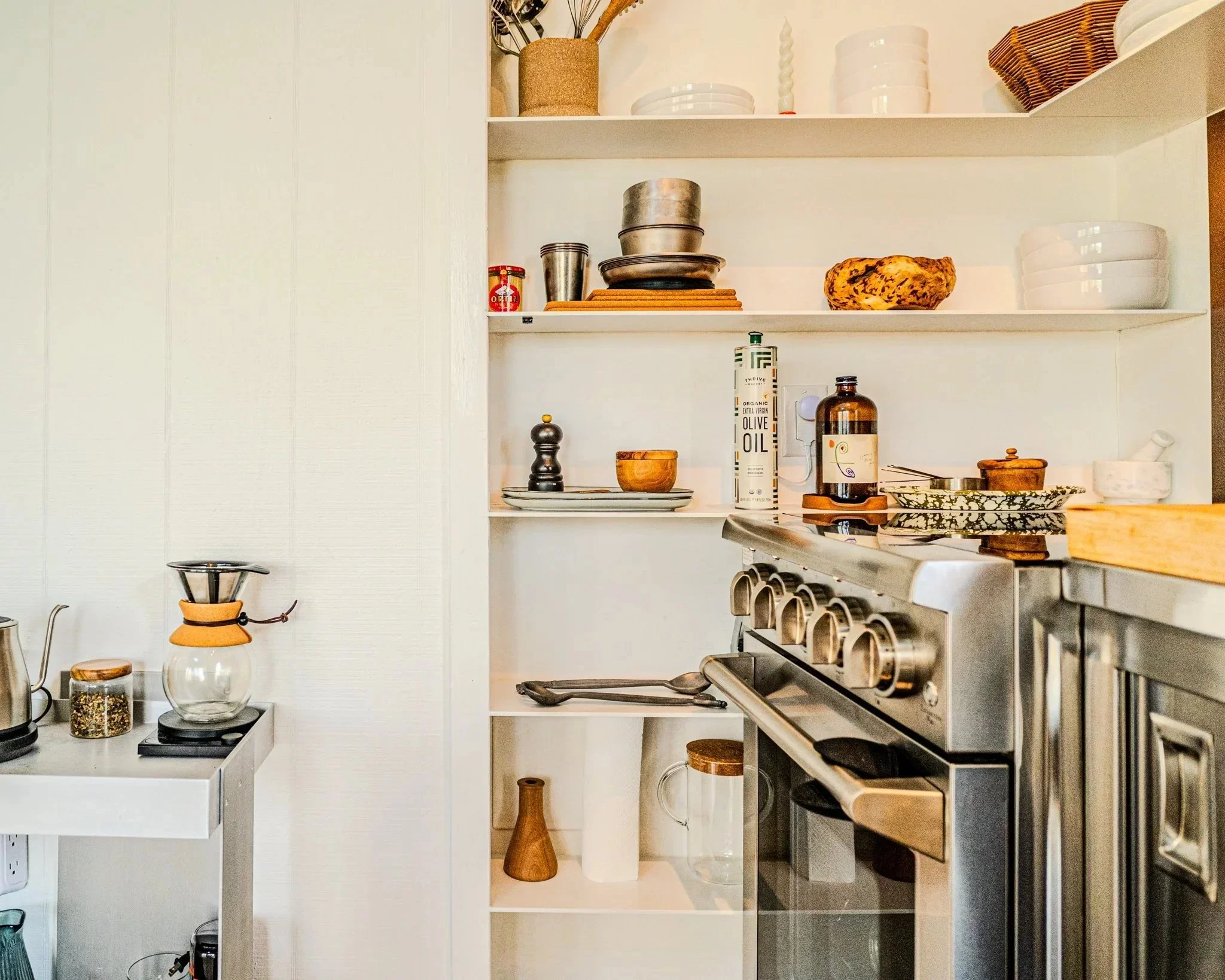 Kitchen shelving and counters with dishes, bowls, aluminum pans, a bread loaf, jars, and a stove with utensils.