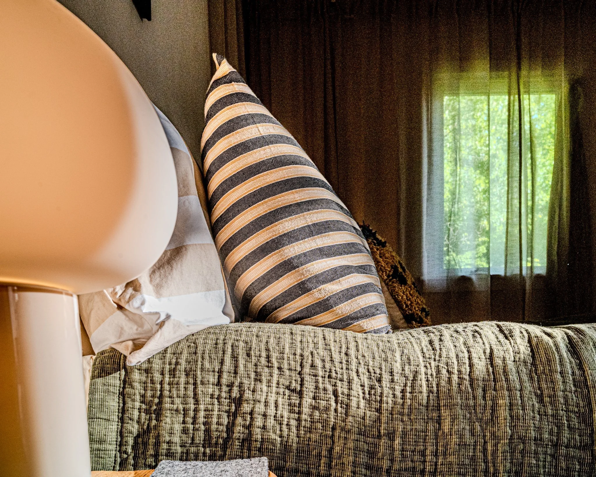 Close-up of a bed with a striped pillow, a blanket, a white lamp on a bedside table, and a window with sheer curtains showing trees outside.