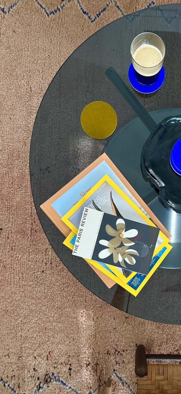 A round glass coffee table with a blue coaster and a glass of beverage, some magazines, a blue pen, a yellow circle coaster, and part of a person's foot visible on a carpet with a patterned design.