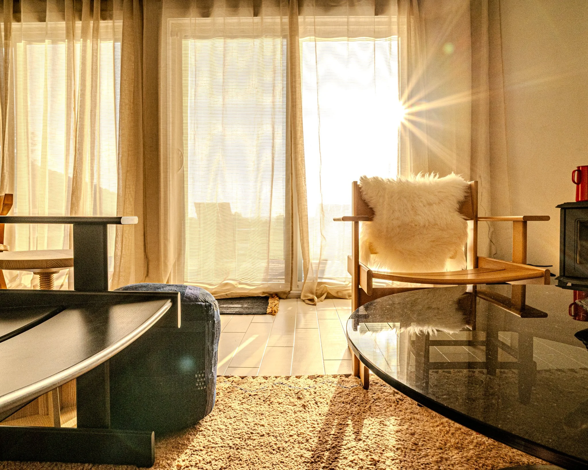 Sunlight streaming through curtains into a cozy living room with a wooden chair and fluffy pillow, a black coffee table, and a beige rug.