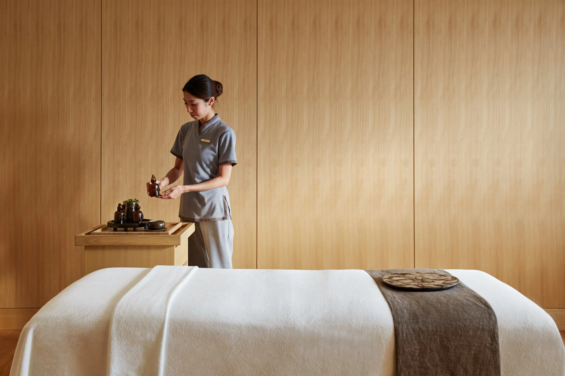 Mitarbeiterin einer Wellness- oder Spa-Einrichtung steht neben einem Behandlungsbett und bereitet ätherische Öle für eine Massage vor.