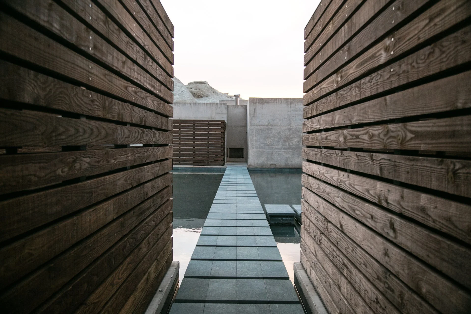 Moderner Außenbereich mit Holz- und Betonwänden, einer schmalen Brücke über Wasser und einem minimalistischen Design.