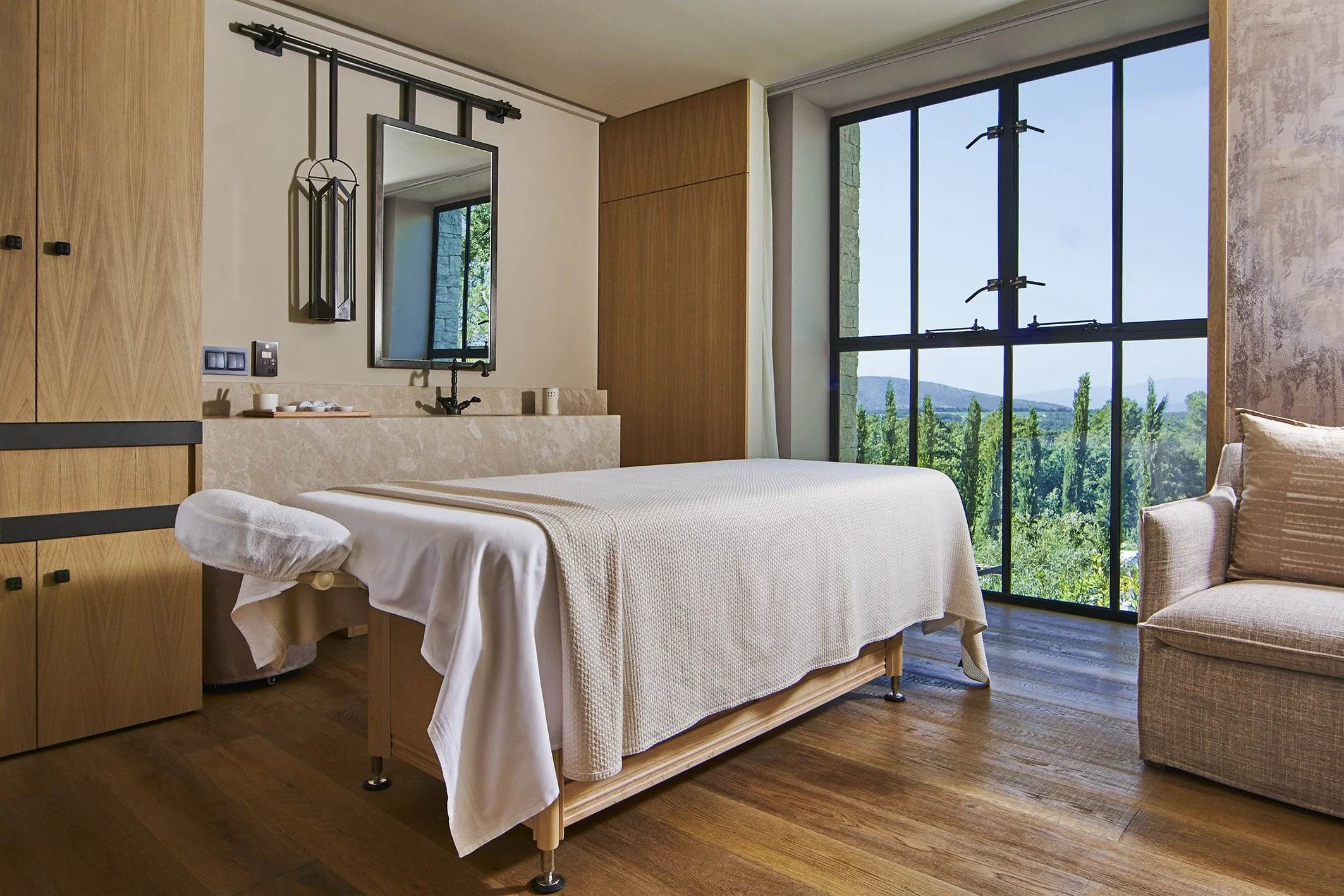 Ein Raummassagebett in einem modernen, hellen Raum mit großen Fenstern und Aussicht auf grüne Bäume und Berge. Der Raum ist mit Holzmöbeln und neutralen Farben gestaltet.