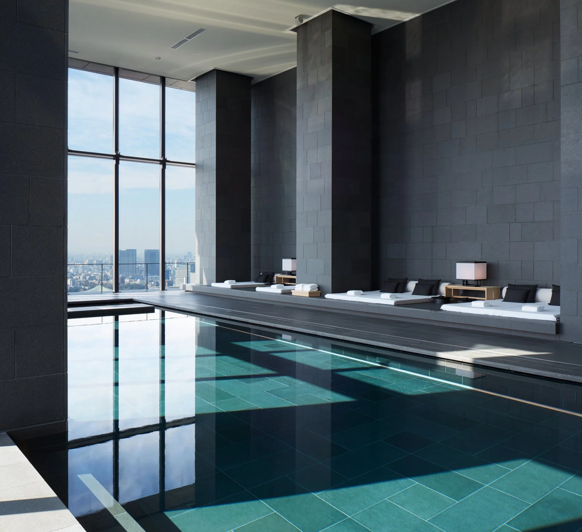 Innenaufnahme eines modernen Wellnessbereichs mit einem Schwimmbecken, Liegen mit weißen und schwarzen Kissen und Seiten Tischen mit Lampen, hohe Fenster mit Blick auf die Stadt. 