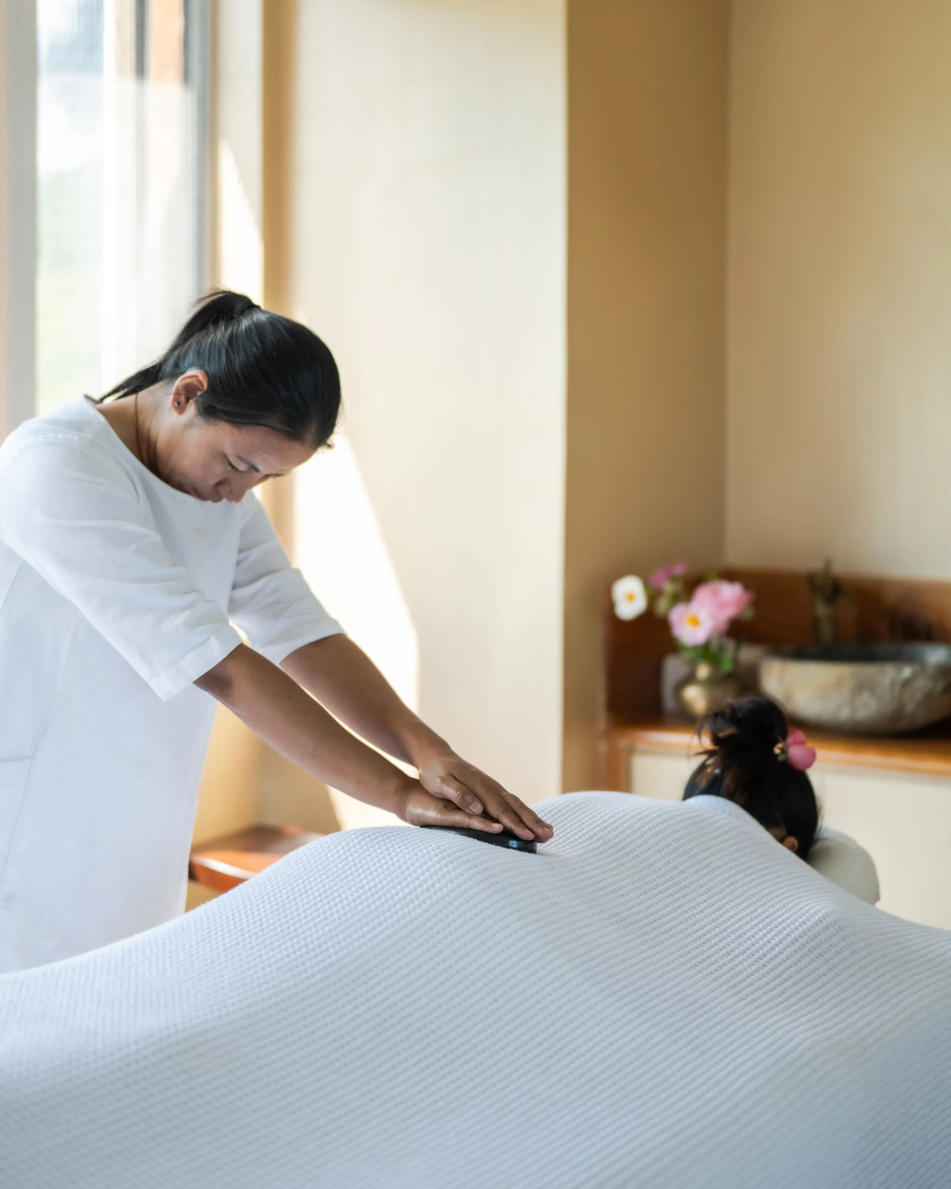 Eine Masseurin gibt einer Person eine Rückenmassage in einem Raum mit natürlichem Licht und Blumen im Hintergrund.