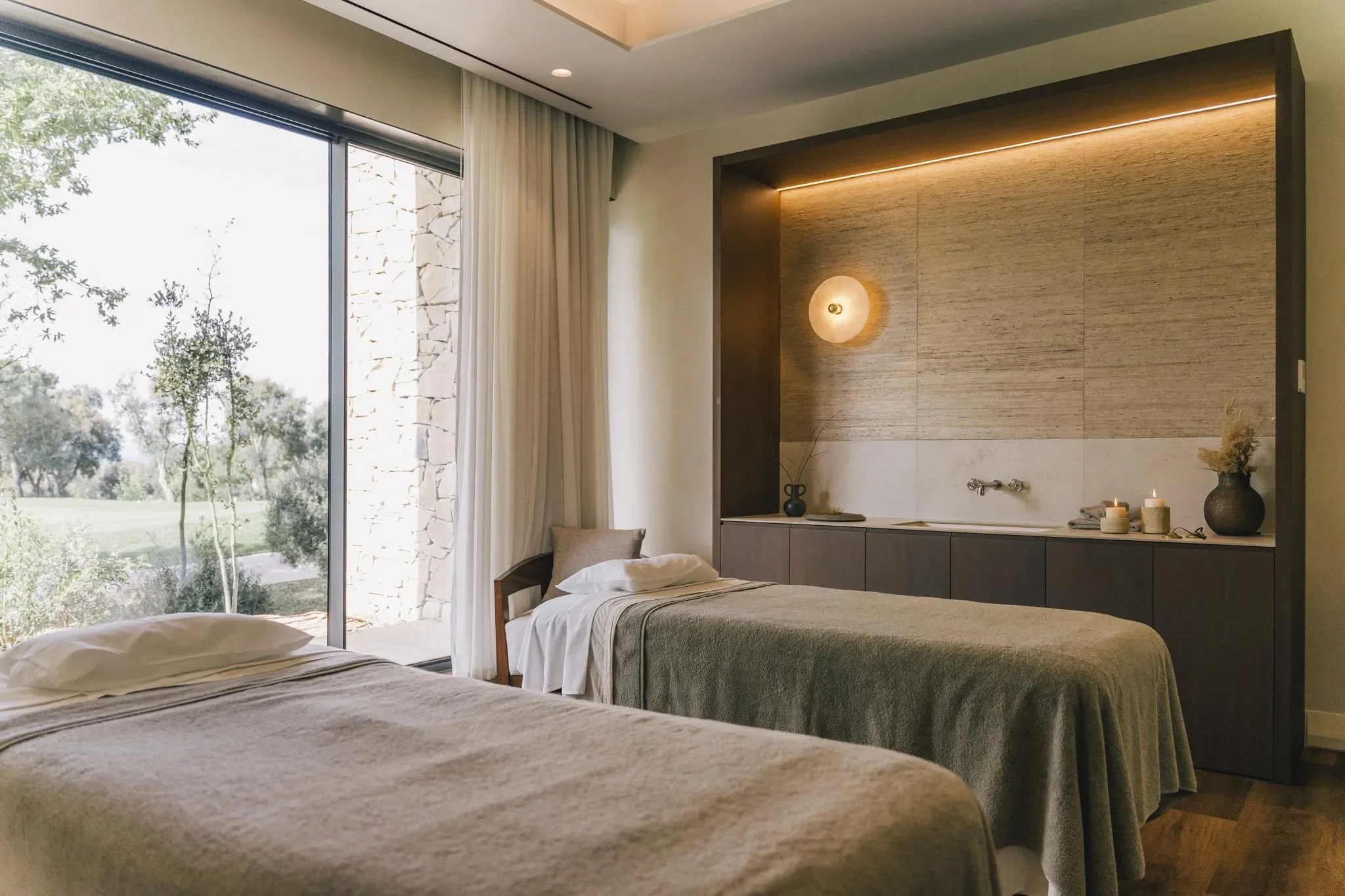 Ein Zimmer mit zwei Betten, einem großen Fenster mit Blick ins Freie, einer Wandlampe und Dekorationsgegenständen auf einer dunklen Kommode.