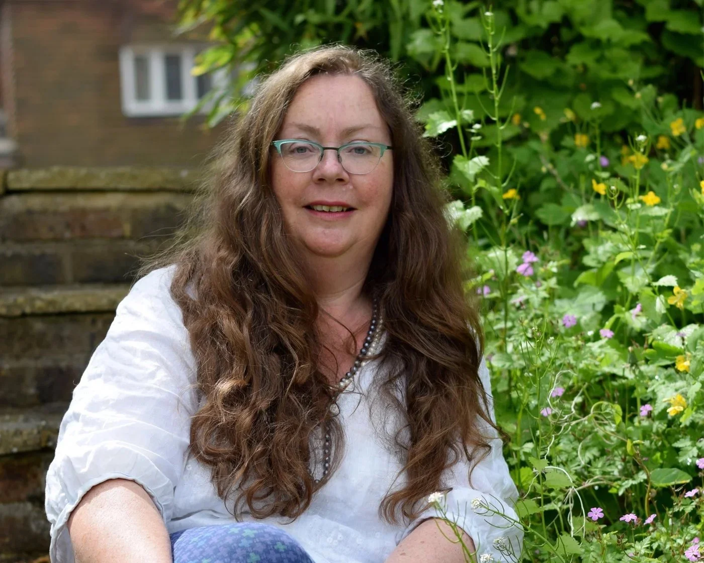 Eine lächelnde Frau mit Brille und langen braunen Haaren sitzt im Garten, umgeben von grünen Pflanzen und bunten kleinen Blumen, mit einer Treppe im Hintergrund.