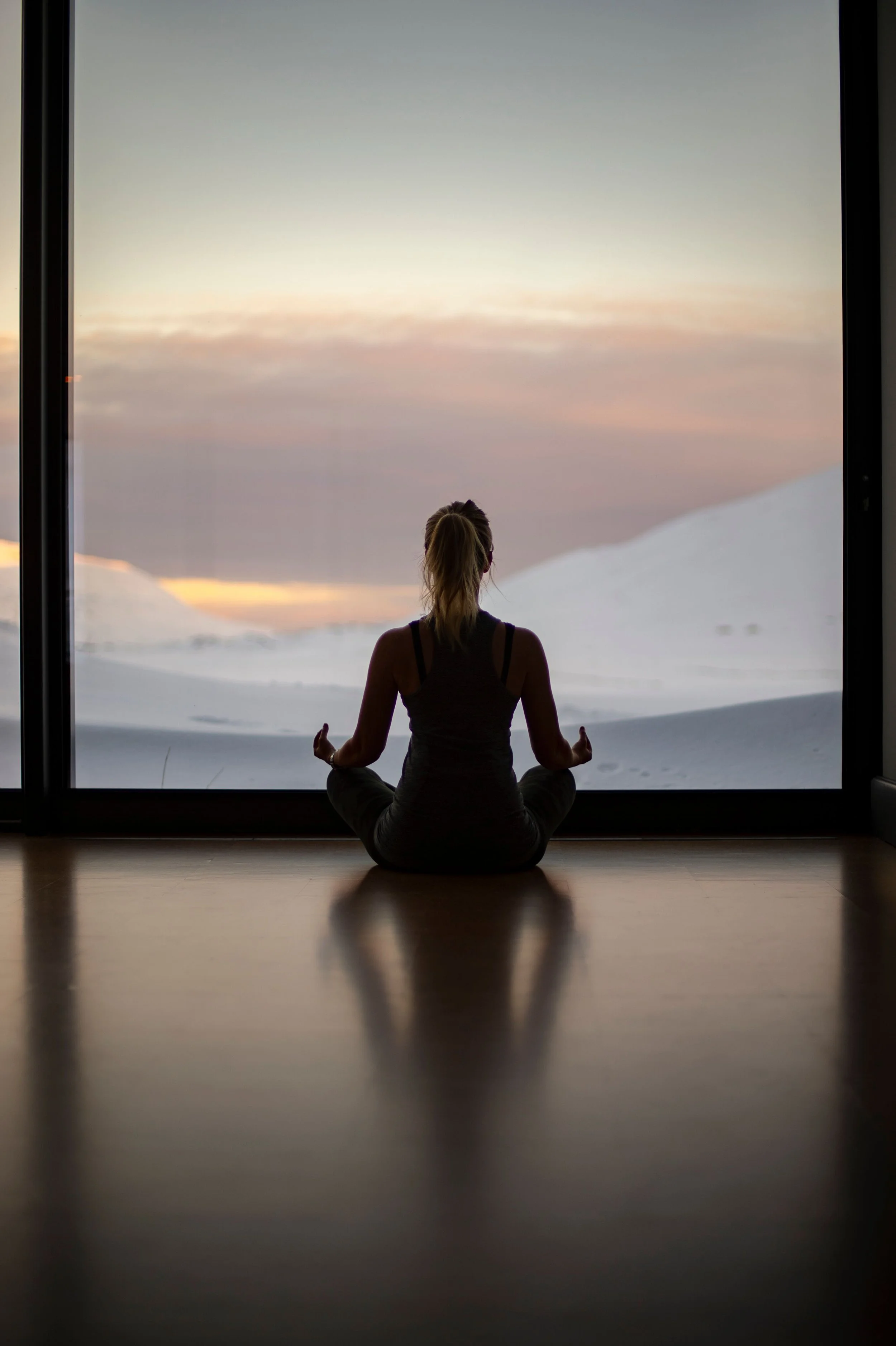 Eine Person in Meditationshaltung sitzt vor einem großen Fenster, das eine schneebedeckte Landschaft mit einem Sonnenuntergang zeigt.