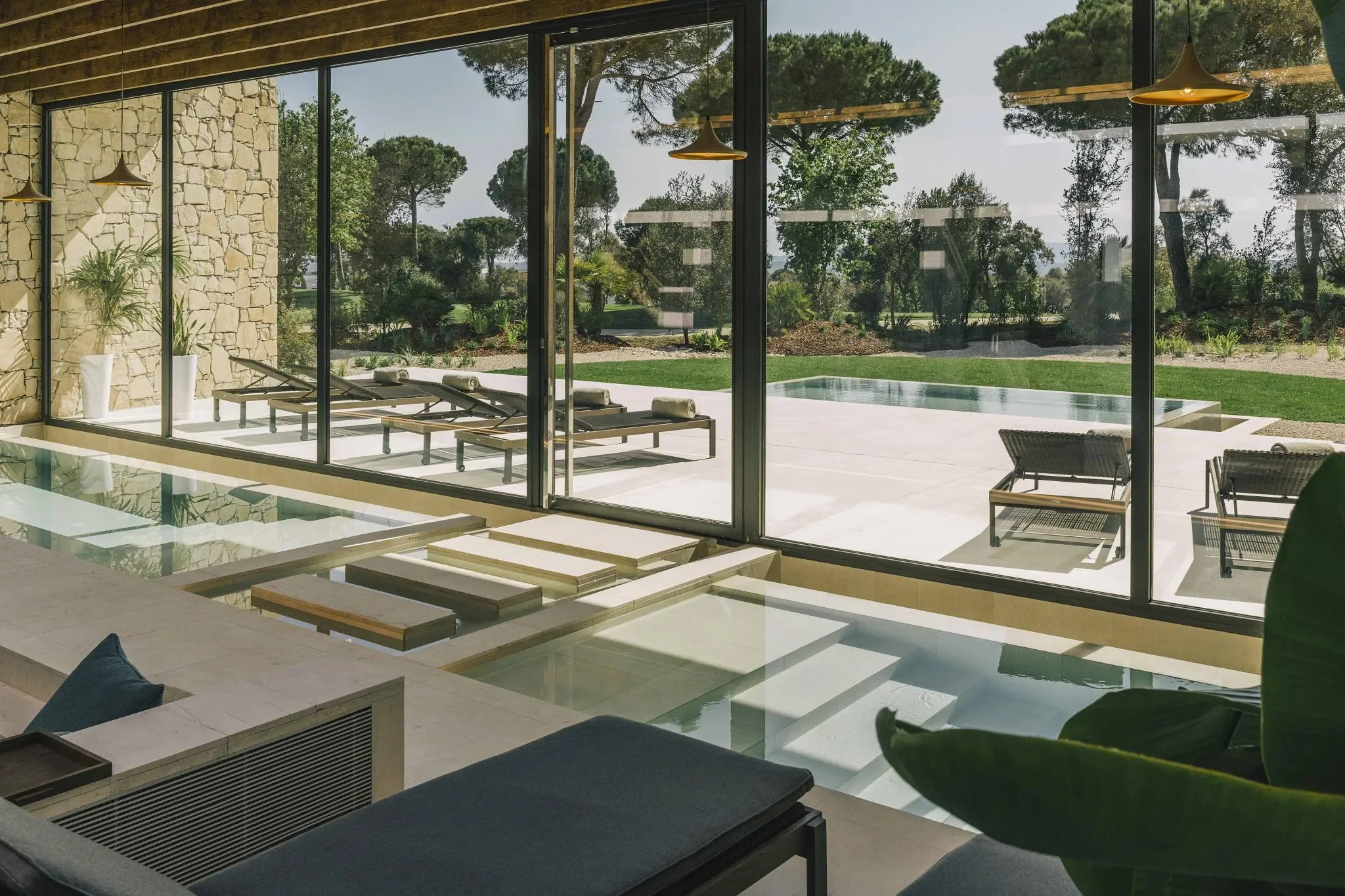 Innenansicht eines modernen Wohnzimmers mit Blick auf den Garten und einen Pool im Freien, große Glasfenster, Liegestühle, Steinelemente an der Wand und bequeme Sitzmöbel.