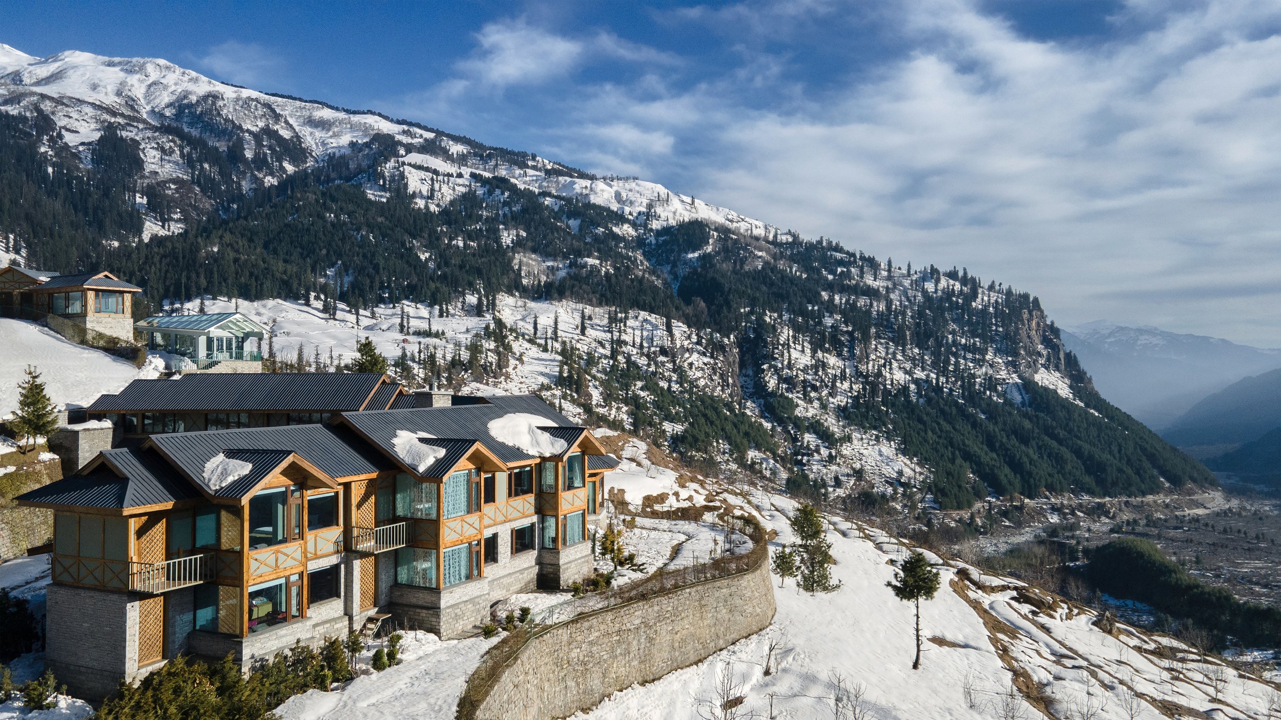 Bergland mit verschneiten Hütten, Bäumen und Bergen im Hintergrund, blauer Himmel mit wenigen Wolken.