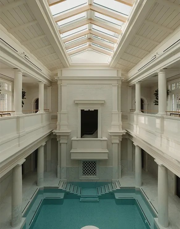 Innenraum eines opulenten, weißen Gebäudes mit einem Pool im Erdgeschoss, umgeben von Säulen und Balustraden, mit großen Fenstern und einem Glasdach.