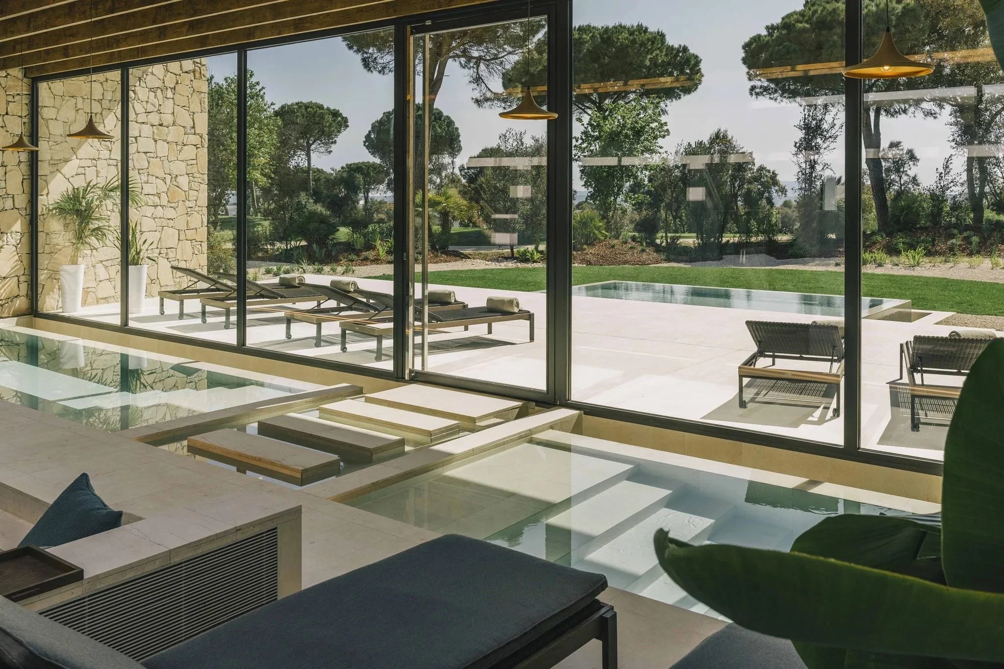 Innenraum mit Blick auf den Garten und den Pool durch große Glasfenster, Liegestühle auf der Terrasse, moderne Einrichtung, Natur- und Steinelemente im Design.