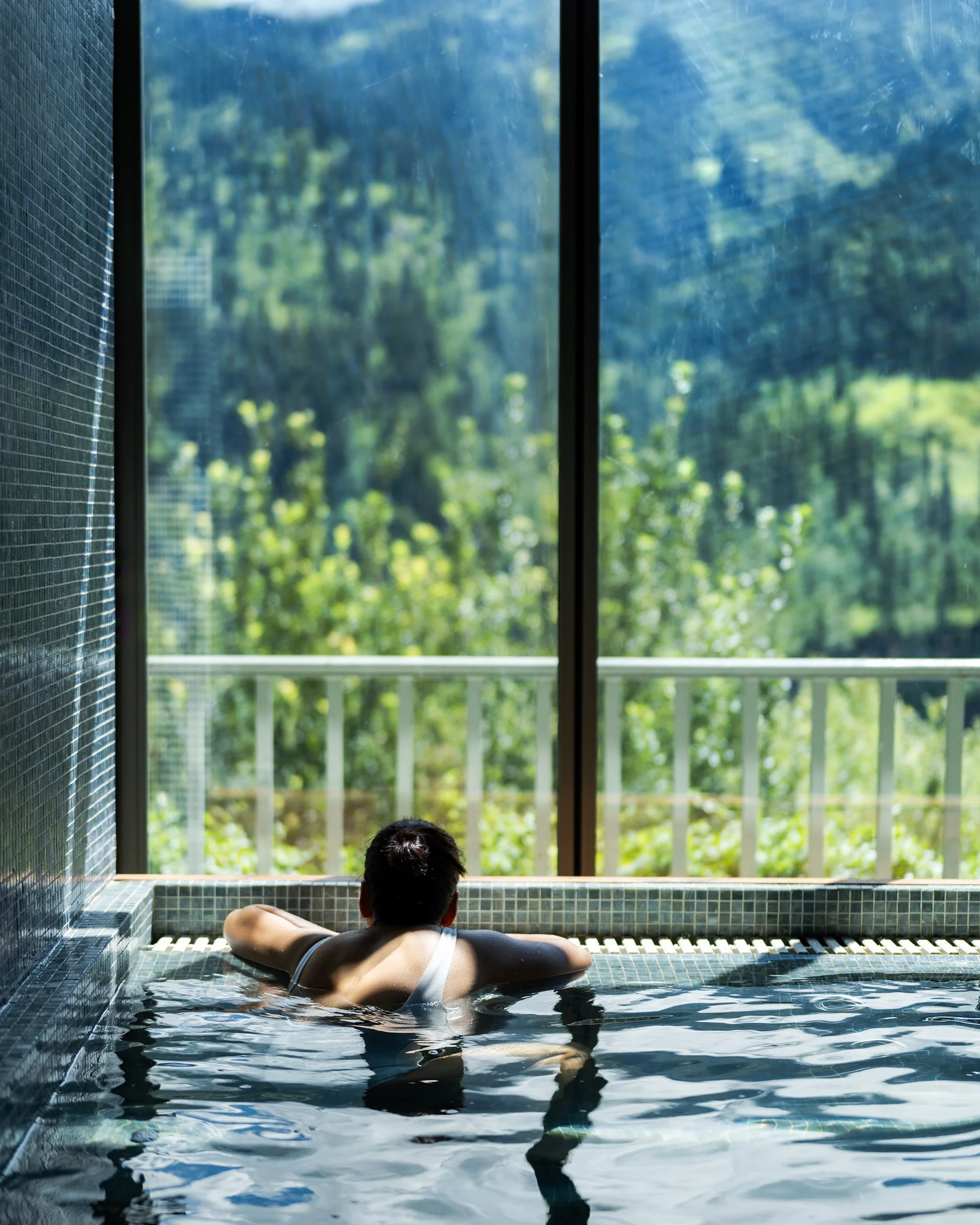 Eine Person liegt entspannt in einem Schwimmbad oder Spa, blickt nach draußen auf eine grüne, bewaldete Landschaft durch große Fenster.