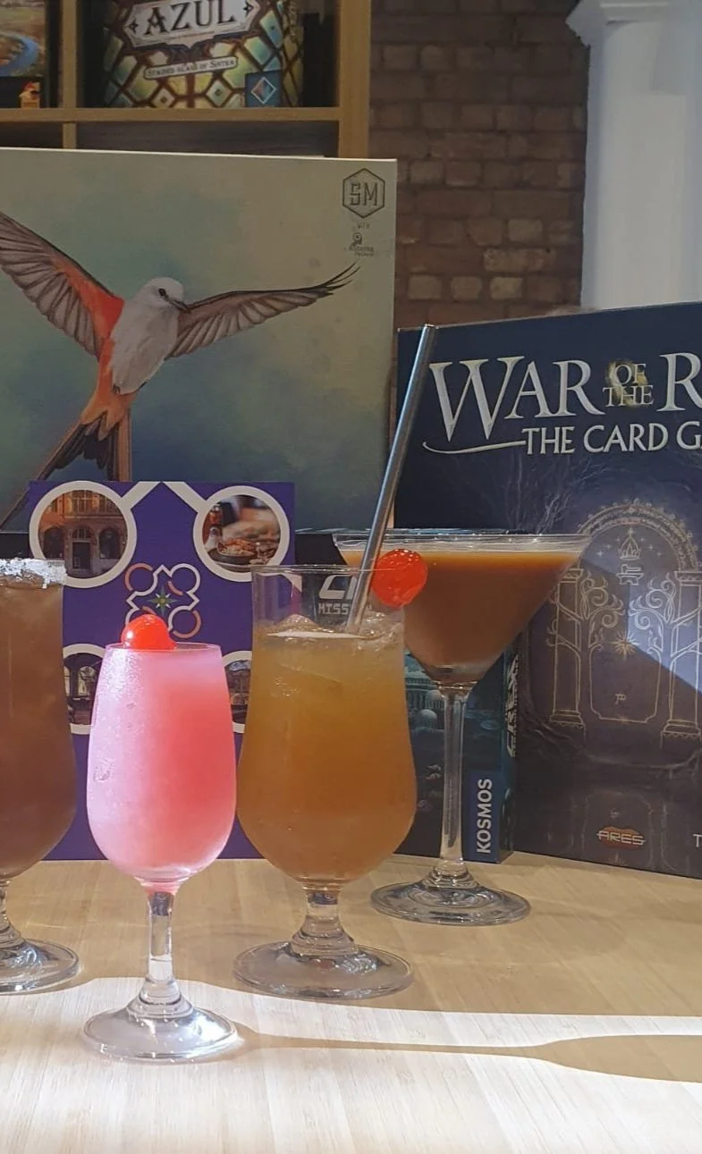 A row of four colorful cocktails on a wooden table, with board game books and artwork in the background.