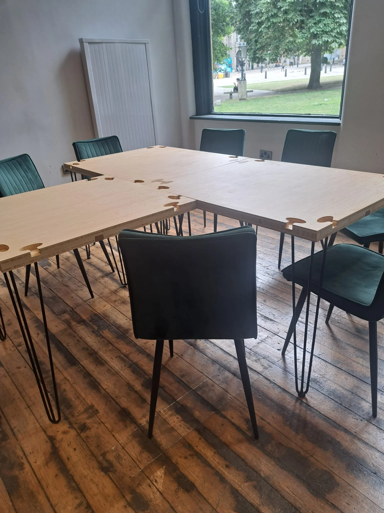 A modern meeting room with a wooden table assembled from puzzle-like pieces, six teal chairs, and a large window showing an outdoor view with trees and the square in front of Sheffield Cathedral.