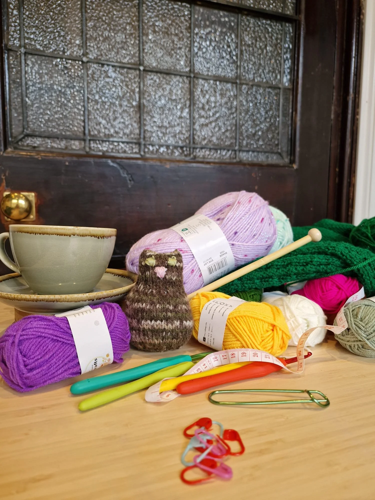 This image shows a hot drink with fibre art tools such as crochet hooks, knitting needles, yarn, etc. It is on a wooden table in a sunny spot in the Gamers Guide Café. It also has a handknitted little kitten plushie in the middle.