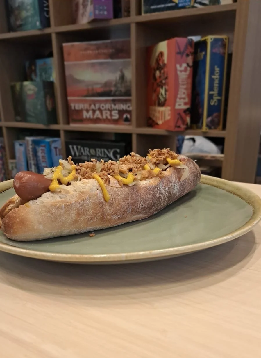 A hot dog on a green plate with mustard and crushed topping, with a background of bookshelves filled with board games.