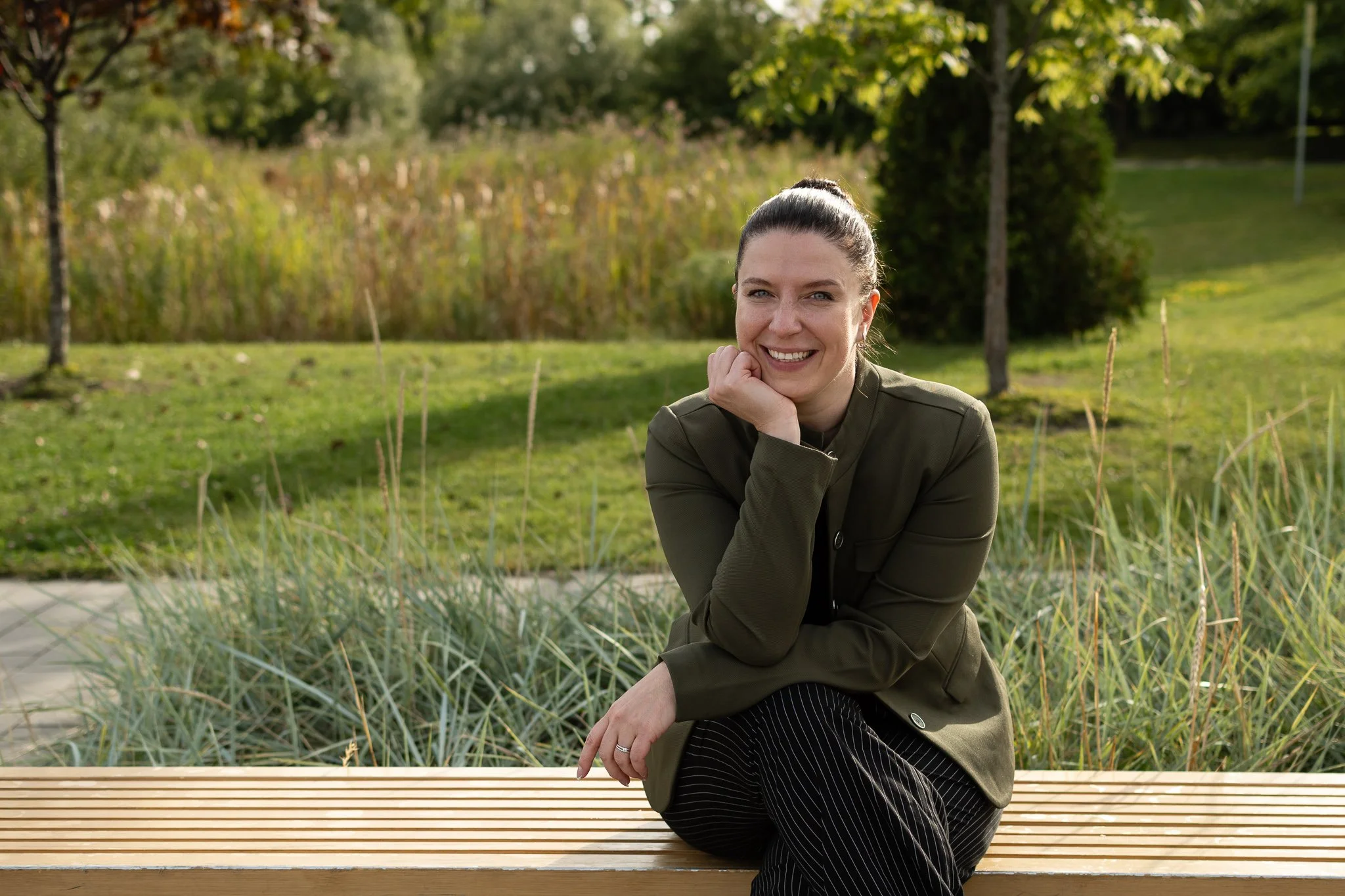 Une femme souriante assise sur un banc en bois dans un parc avec des arbres et de l'herbe en arrière-plan.
