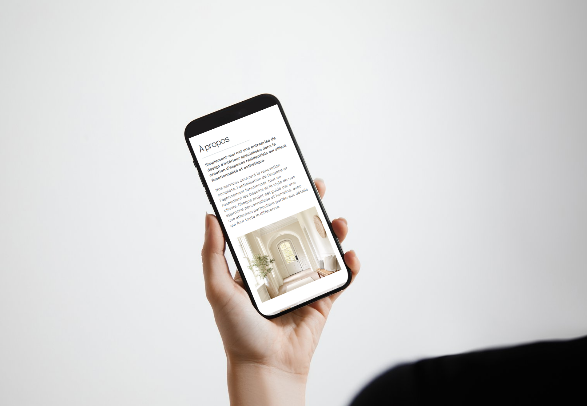 Une main tenant un smartphone avec une page web affichée, contenant du texte en français et une image d'une entrée lumineuse et élégante d'une maison ou d'un espace intérieur.