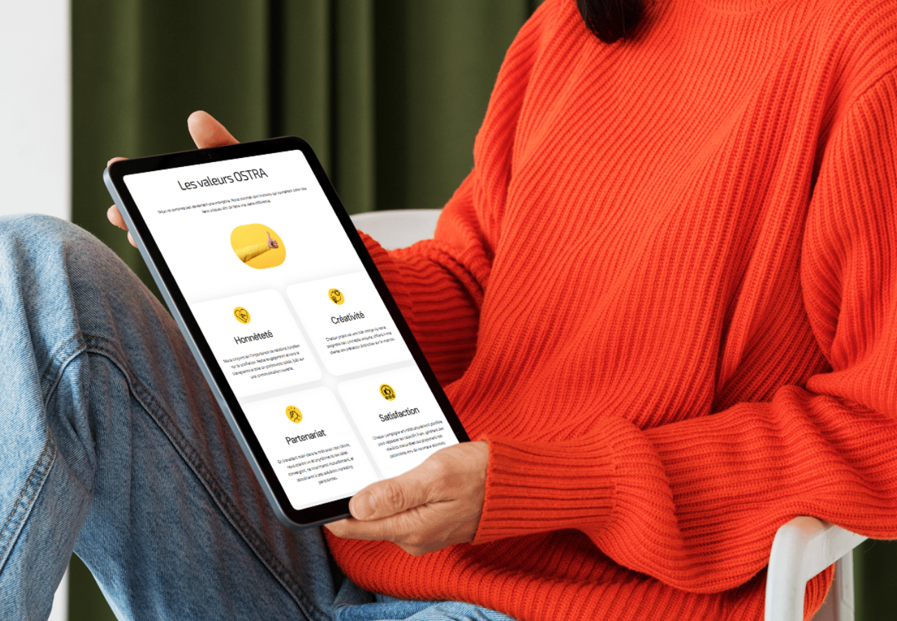 Personne tenant une tablette montrant une application avec des valeurs OSTORA, portant un pull orange, assise sur une chaise blanche.
