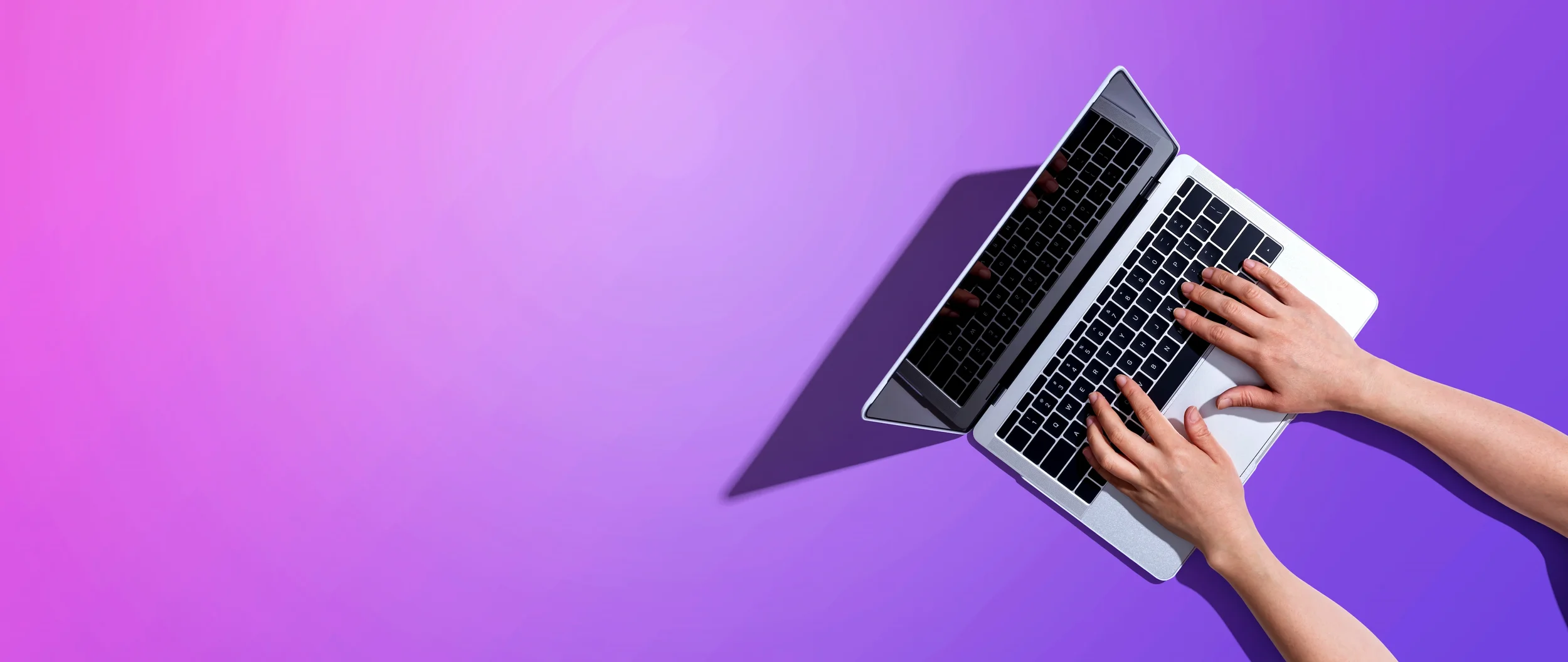 Deux mains tapent sur un clavier d'ordinateur portable argenté placé sur un fond violet