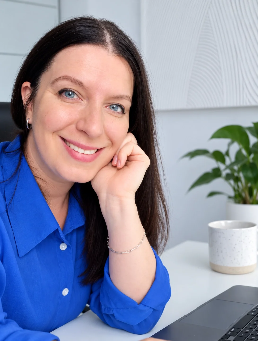 Femme souriante avec cheveux bruns, portant une chemise bleue, assise devant un ordinateur portable dans un bureau moderne avec une plante verte et un tableau blanc en arrière-plan.