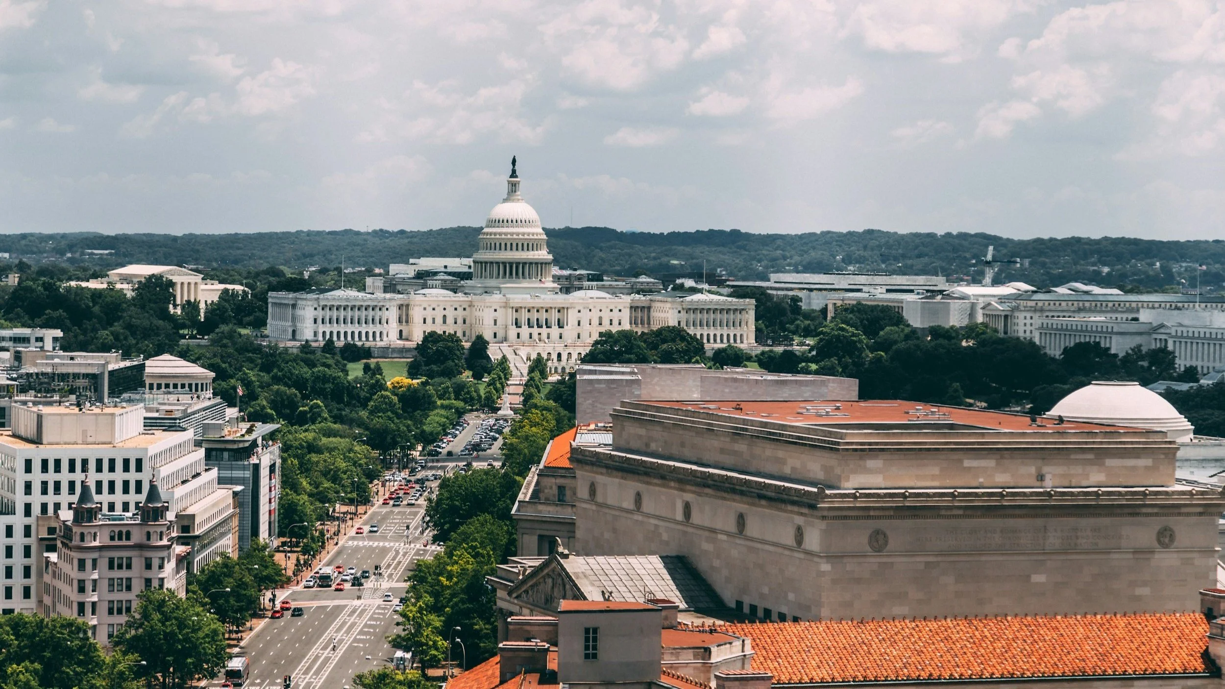 A cityscape featuring the U.S. Capitol building in the background with modern and historic buildings in the foreground, tree-lined streets, and a partly cloudy sky.