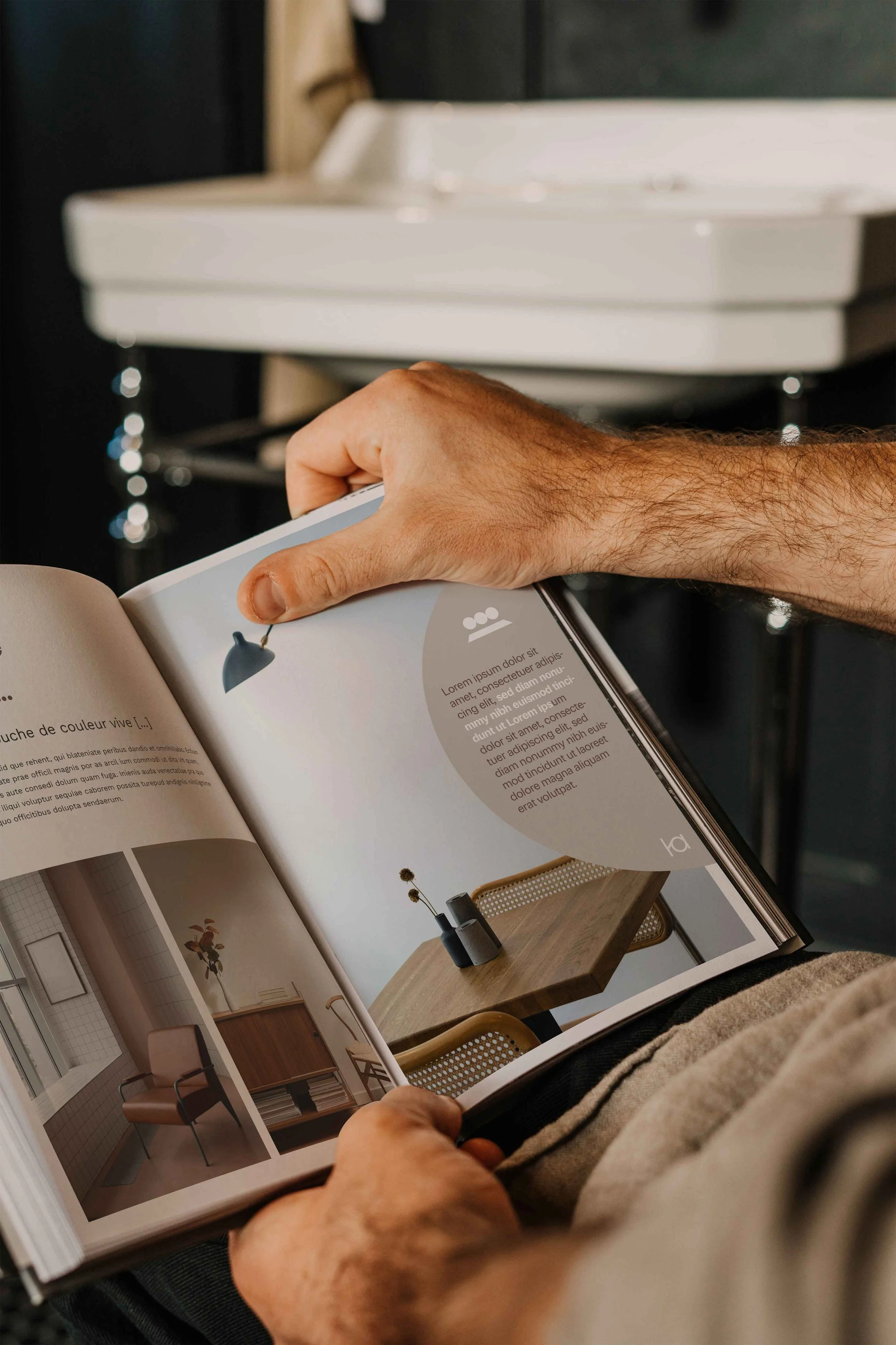 Une personne tient un catalogue d'architecture d'intérieur ouvert en montrant des images de décoration intérieure, notamment un meuble en bois avec un vase et des fleurs.