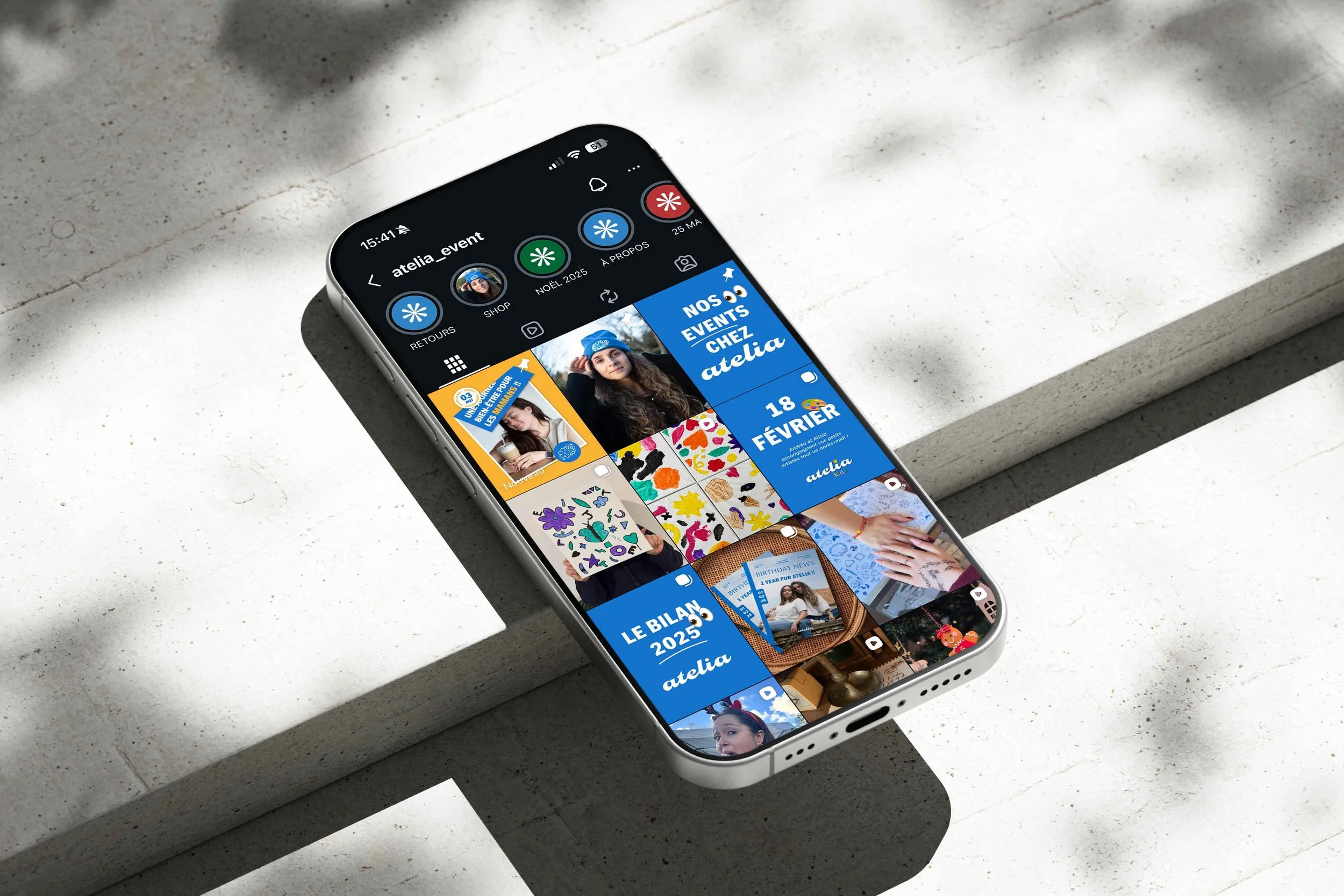 Un smartphone affichant le profil Instagram de atelia_event sur une surface en béton, avec plusieurs stories et publications, notamment des annonces d'événements et des images de créations artistiques.