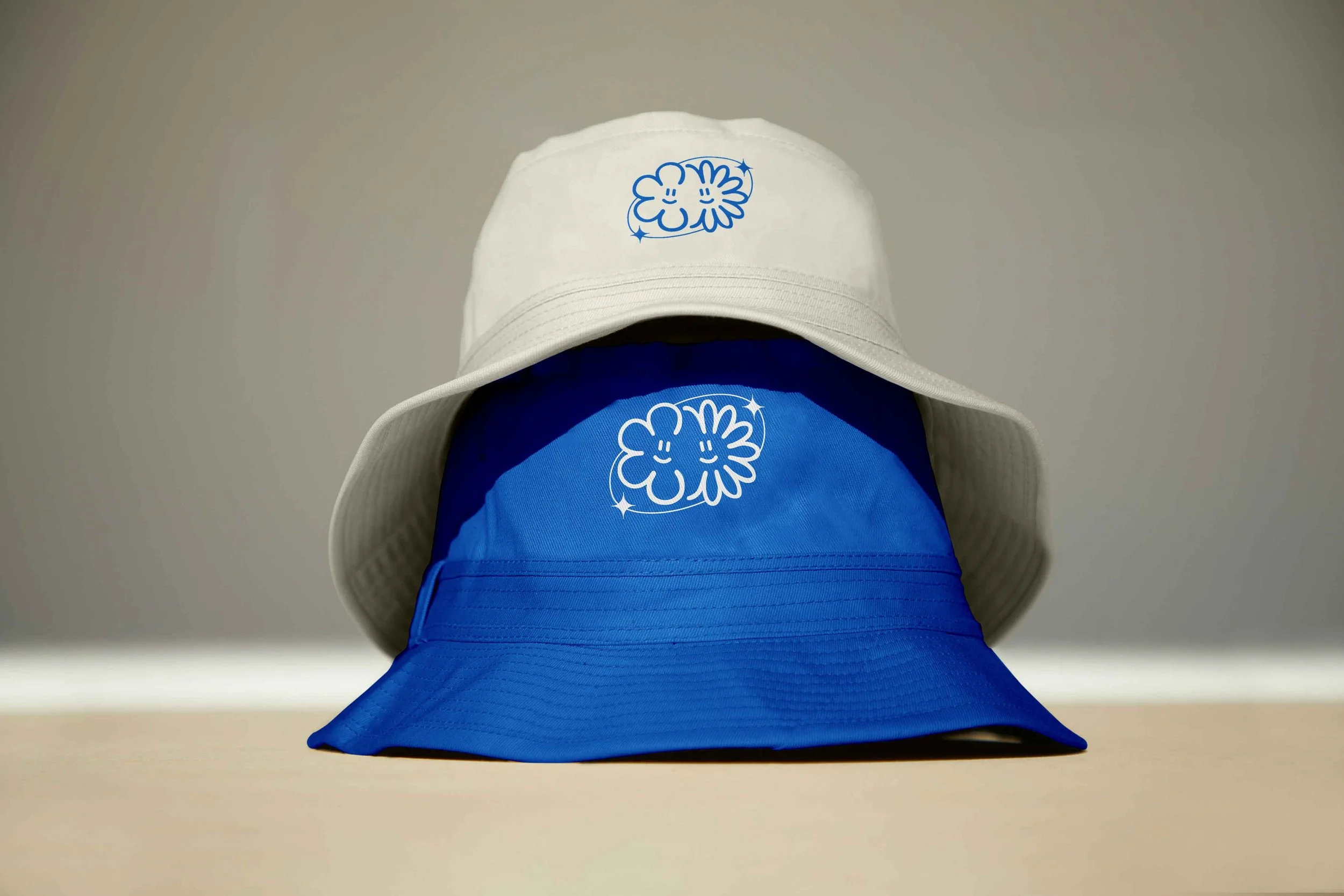Deux chapeaux (bob), un blanc et un bleu, avec le logo d'une association d'événementiel (forme de fleur)