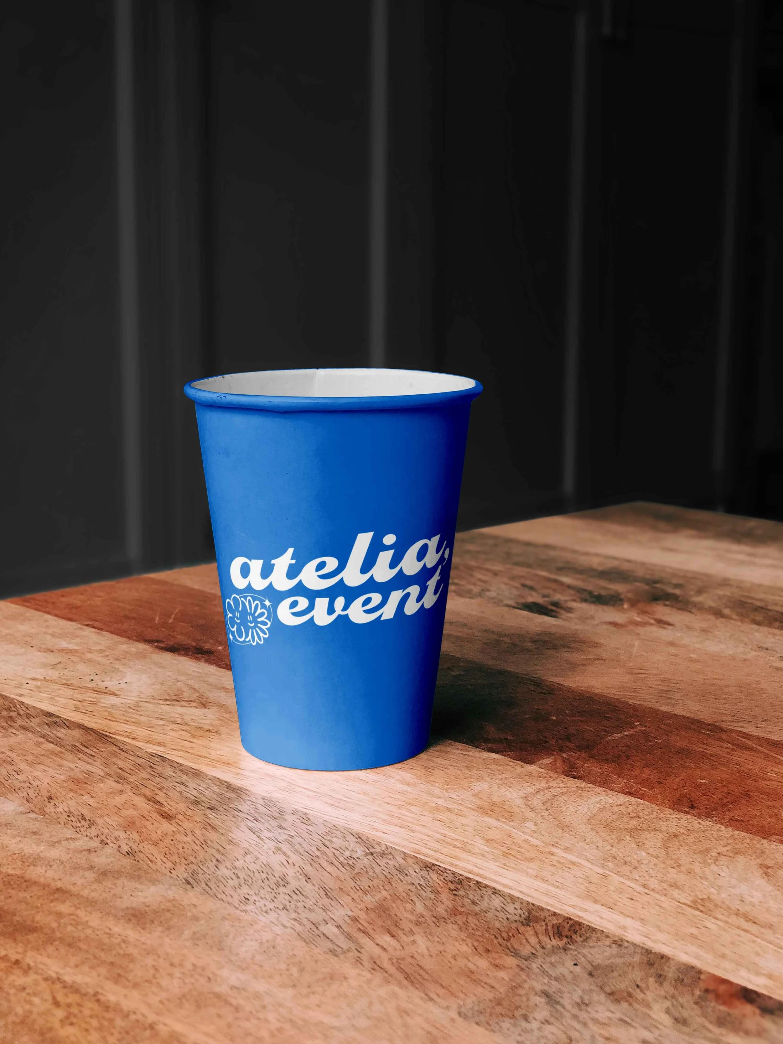 Gobelet en carton bleu avec le texte 'ATELIA EVENT' blanc et avec le logo d'une association d'événementiel (forme de fleur, illustration)