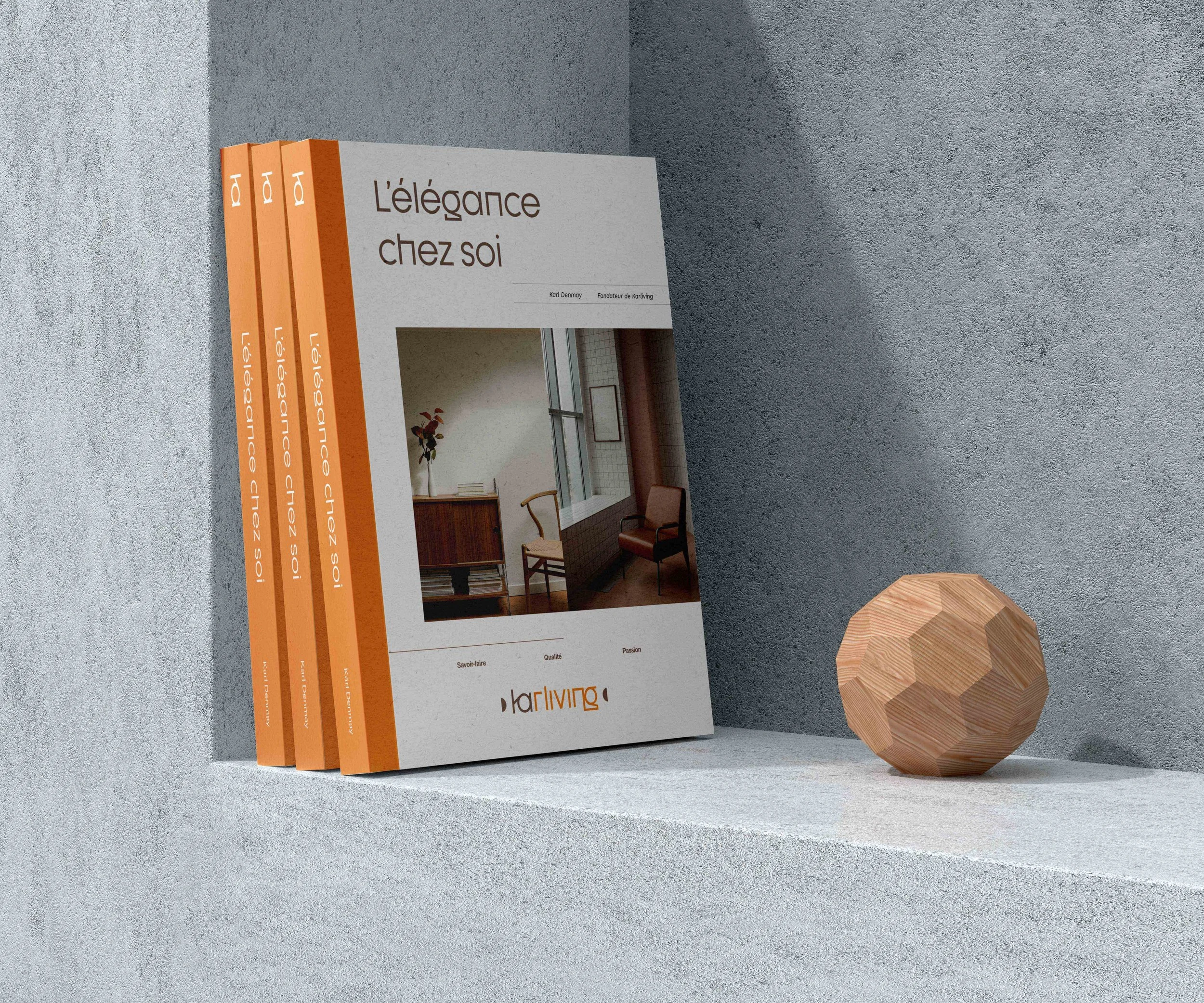 Trois catalogues avec la couverture orange et blanc, intitulés 'L'élégance chez soi', se tenant contre un mur gris et une surface blanche, à côté d'une sculpture géométrique en bois