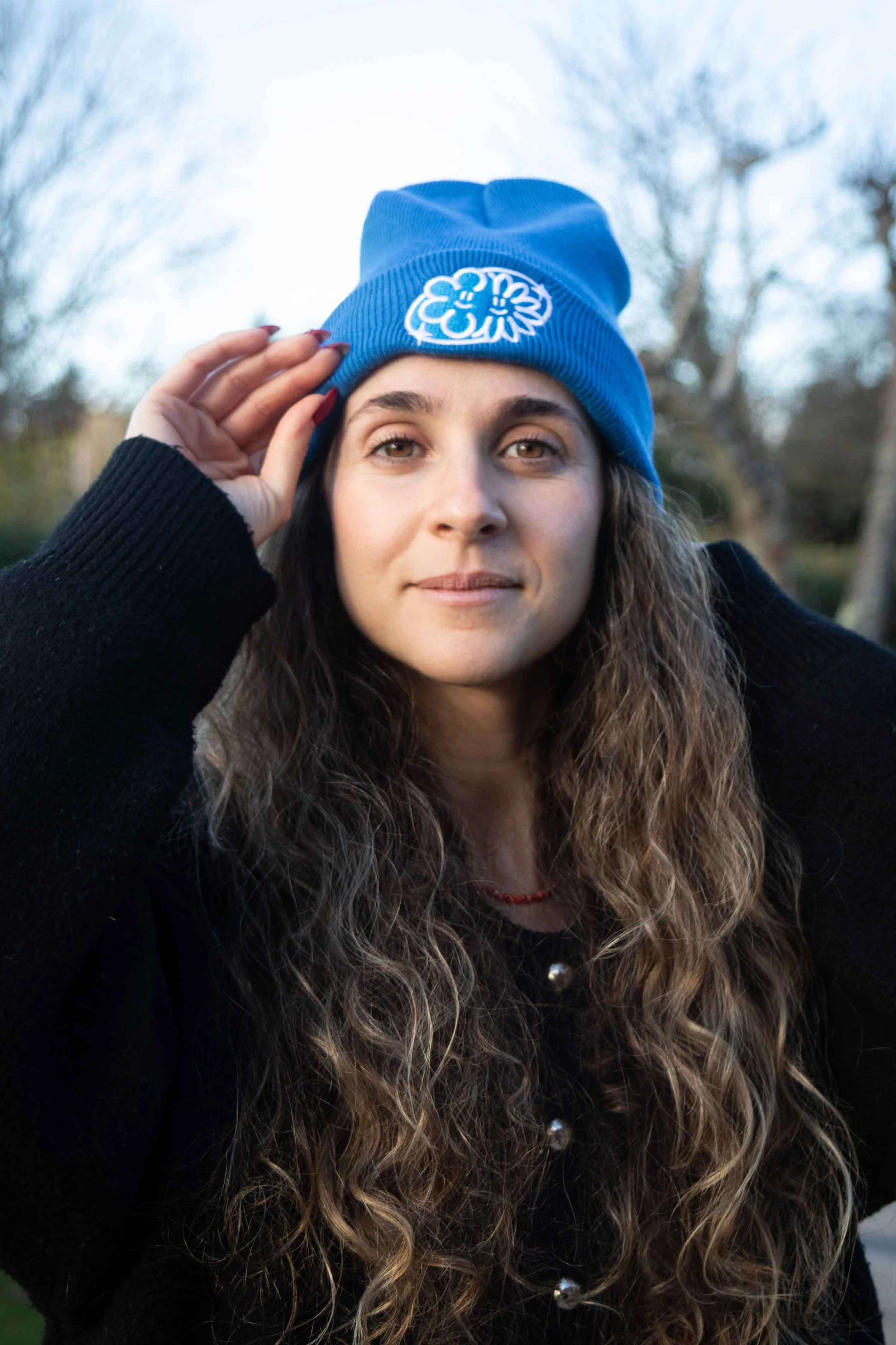 Jeune femme avec cheveux ondulés, portant un bonnet bleu avec un logo blanc, et un pull noir, à l'extérieur avec des arbres en arrière-plan.