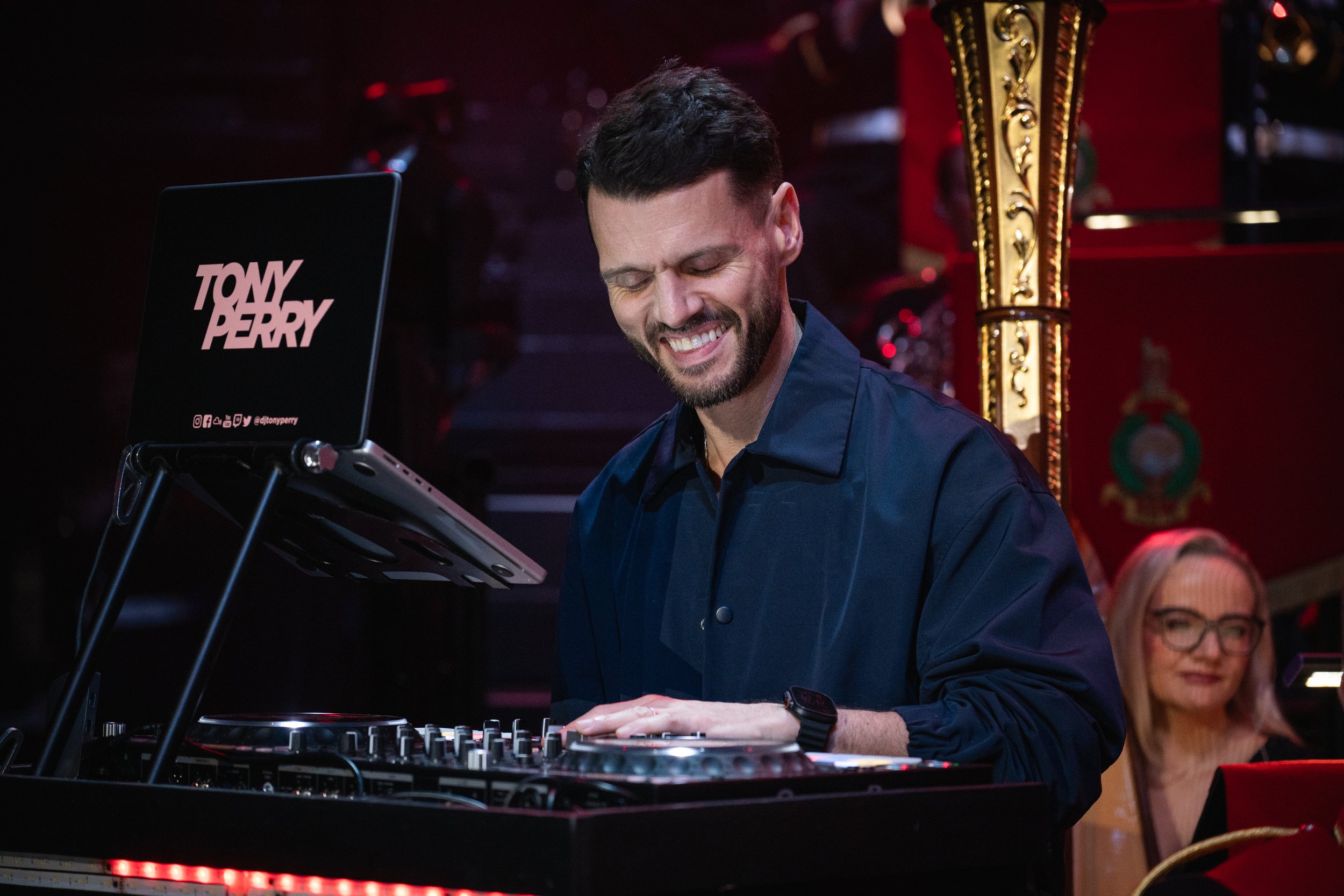 A man with dark hair and a beard smiling while DJing at an event, wearing a dark blue shirt and a watch, with a DJ controller in front of him. There are people in the background and holiday decorations visible.
